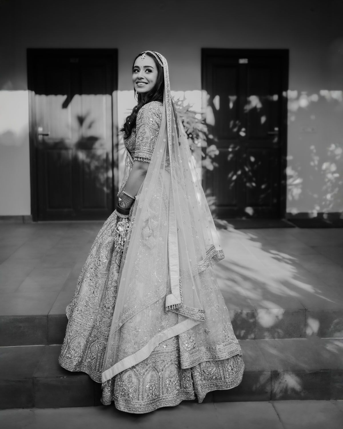 Black and white photograph of a bride wearing an ornate lehenga and flowing veil posing in front of a shadowy backdrop.