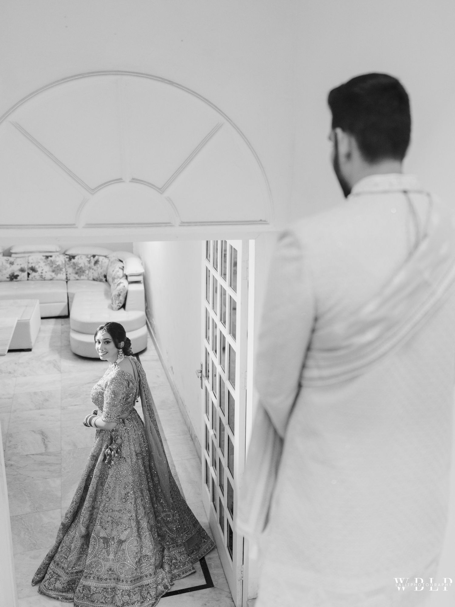 Black and white photo of groom watching bride in elegant lehenga during Indian wedding first look ceremony.