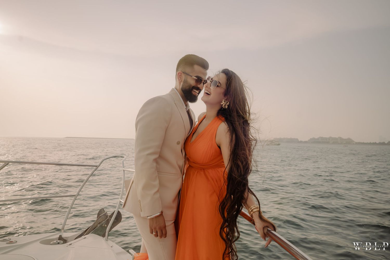 Romantic couple embracing on a boat at sunset, woman in orange dress, man in beige suit, ocean background.