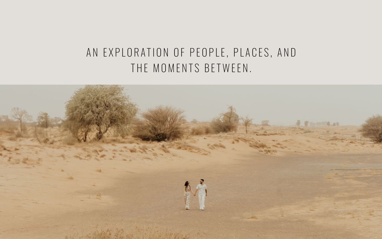 Two people in white attire walk hand-in-hand through a vast desert landscape with sparse trees under a muted sky.