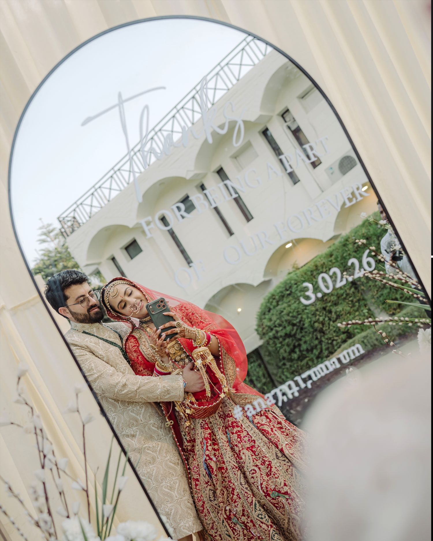 Couple in traditional Indian wedding attire taking a mirror selfie, bride in red lehenga and groom in cream sherwani.