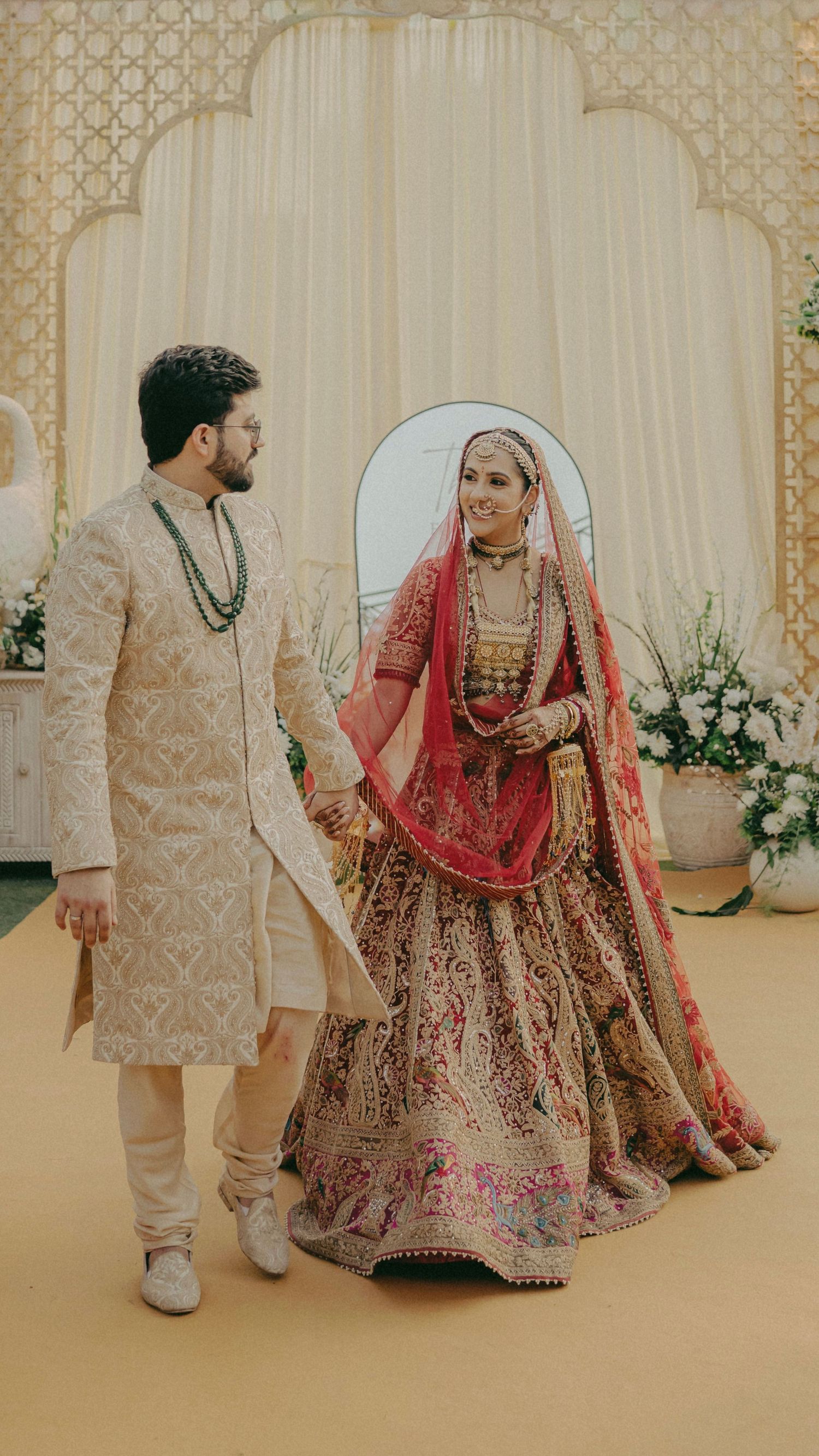 Indian bride in red and gold lehenga holds hands with groom in cream sherwani at elegant wedding ceremony.