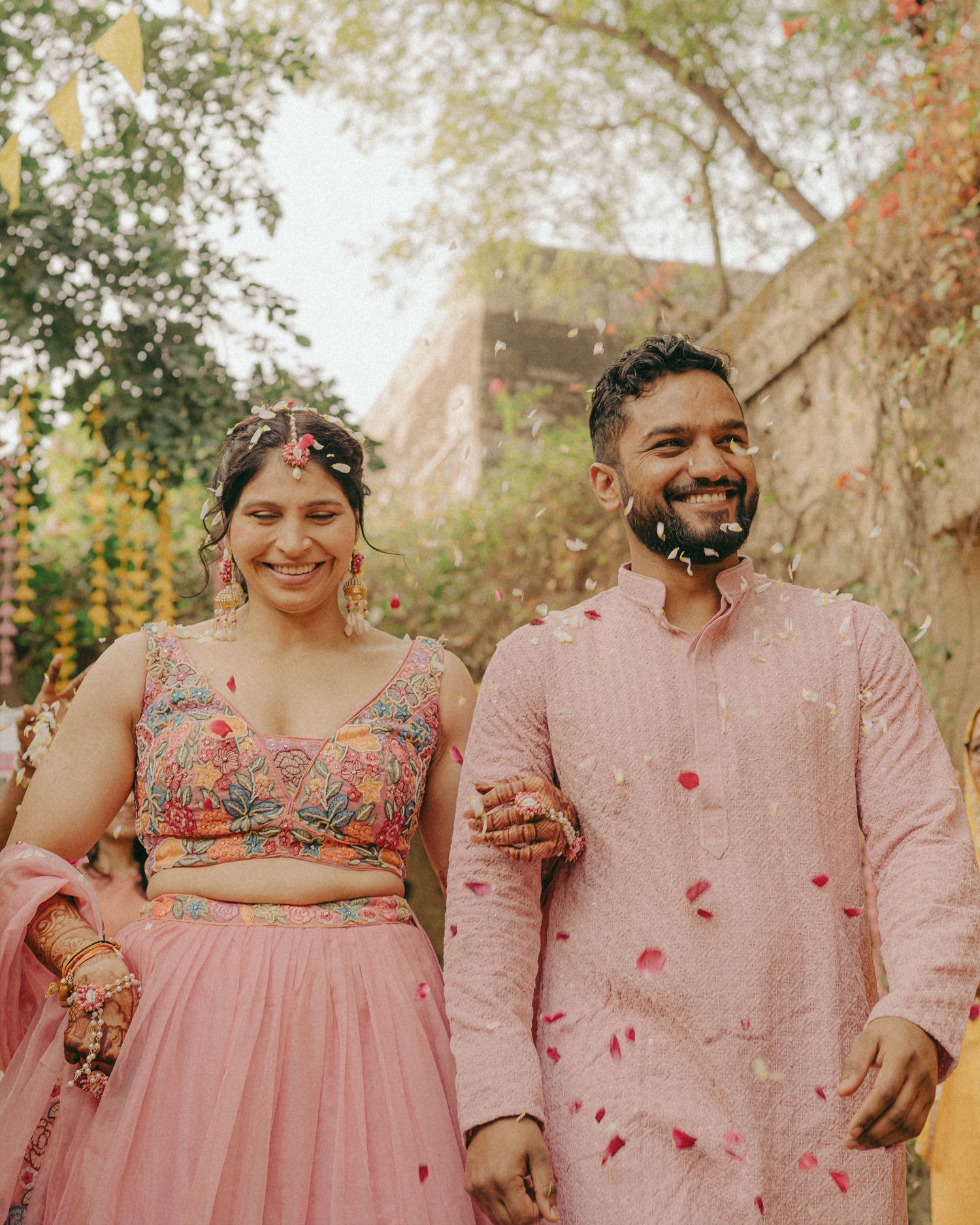 Happy couple in matching pink traditional Indian wedding attire, smiling as flower petals fall around them.