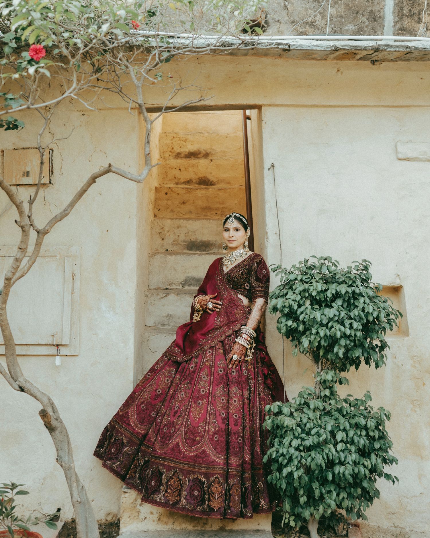 A bride in a stunning dark red and gold embroidered lehenga poses gracefully outside a traditional Indian building.