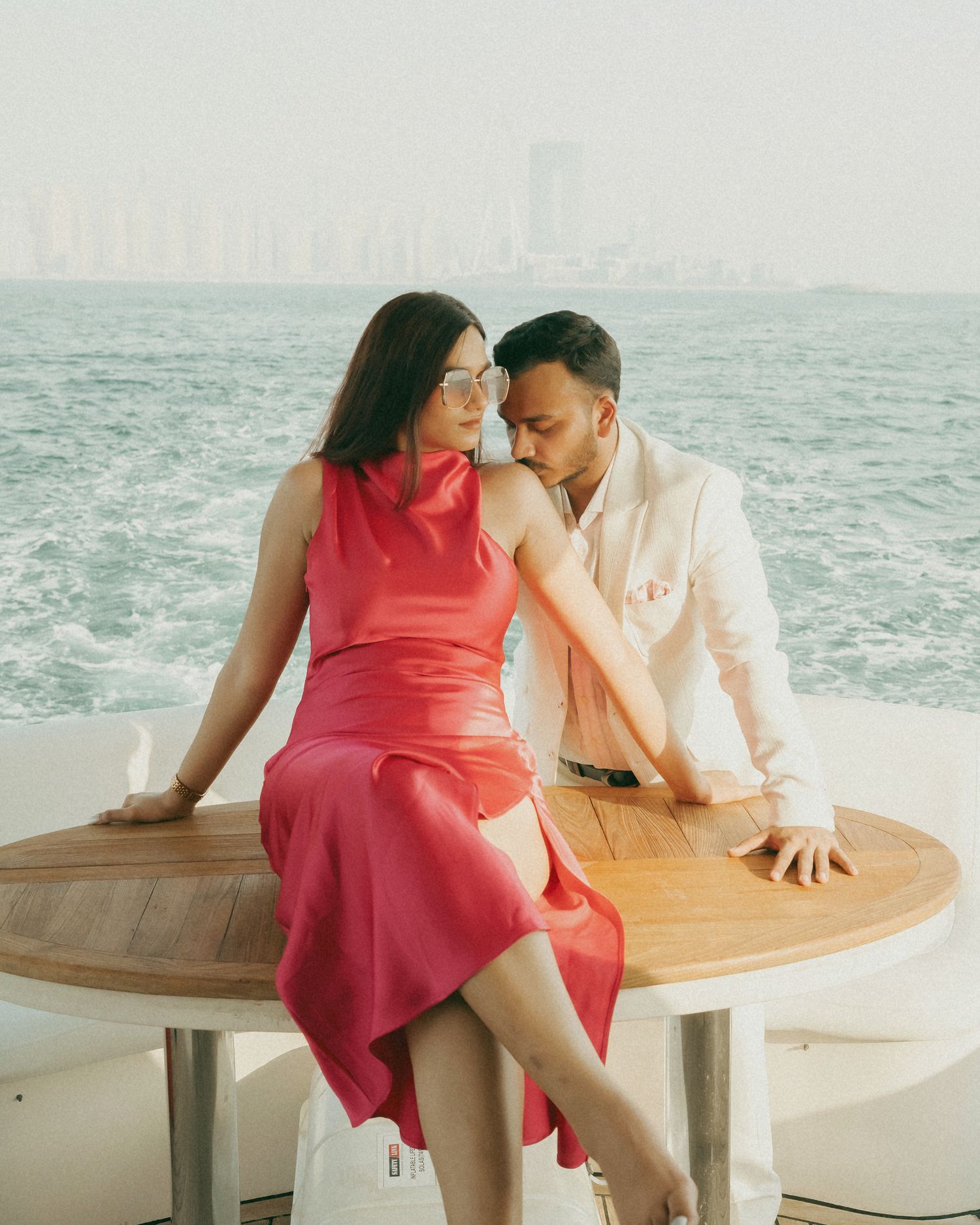 Couple in elegant attire sharing a romantic moment on a yacht with city skyline backdrop.