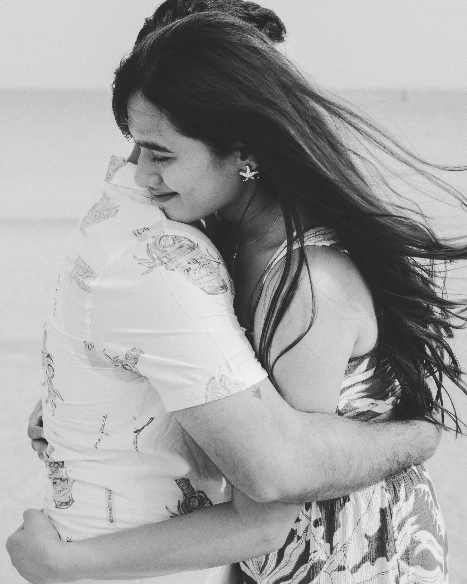 Black and white photo of a couple embracing on a beach, woman with long hair smiling as wind blows.