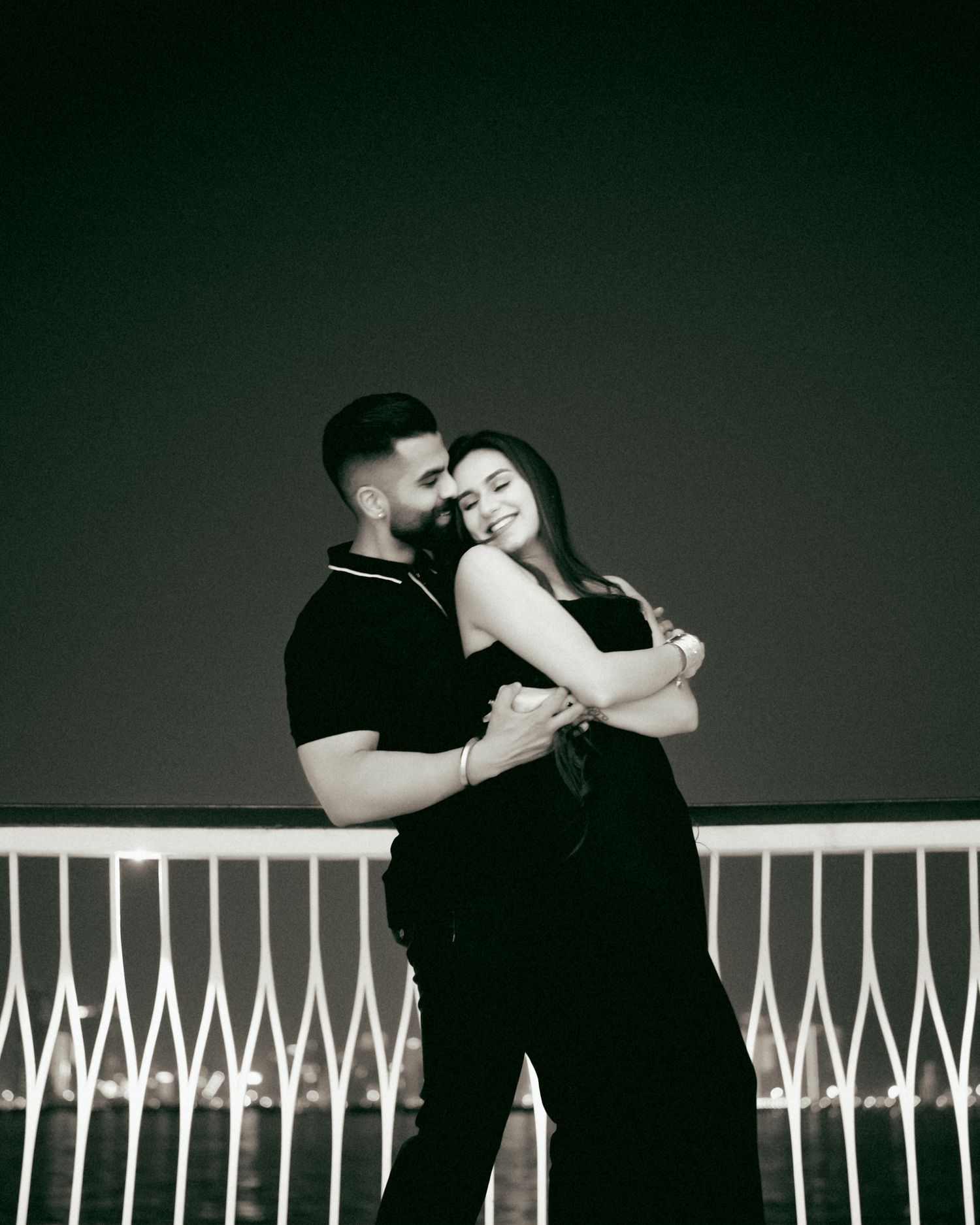 Black and white romantic couple embracing on a balcony at night, woman leaning into mans chest affectionately.