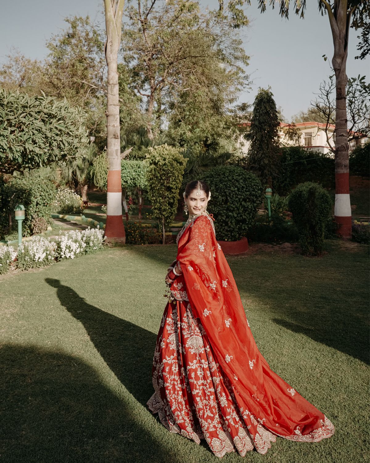 A bride in a flowing red lehenga poses gracefully in a garden setting.