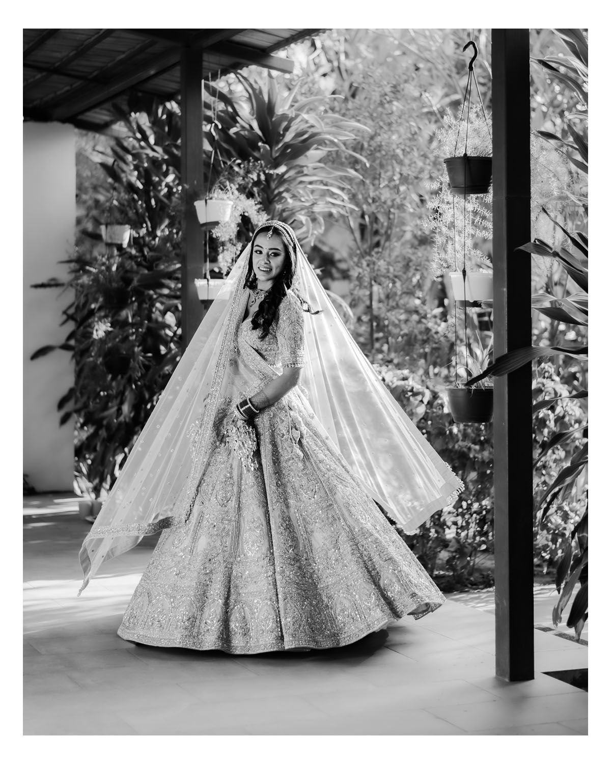 Black and white photo of a bride in a sparkly ballgown wedding dress with long veil posing in a tropical garden setting.
