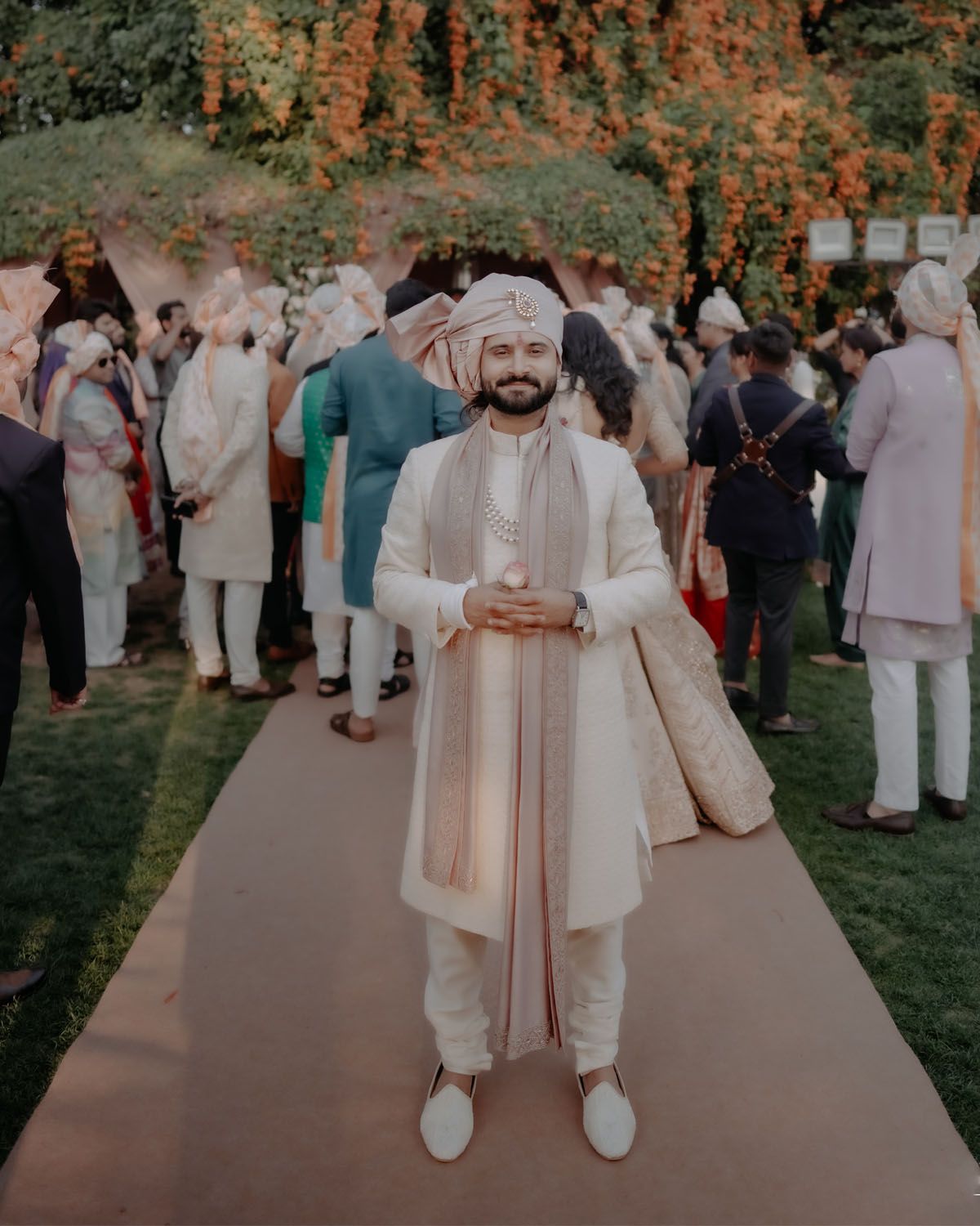 A person wearing traditional white and beige Indian wedding attire stands on a path at an outdoor celebration.