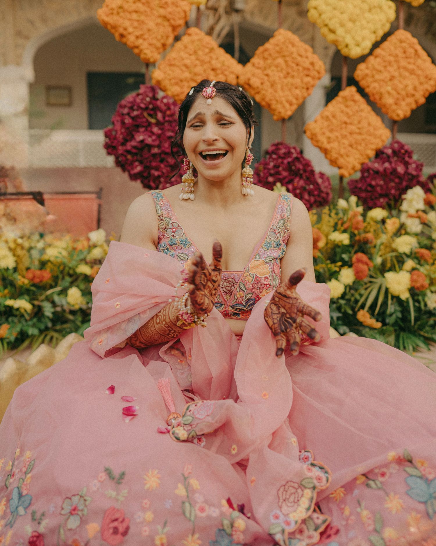Woman in pink floral lehenga with mehndi hands at Indian wedding ceremony with colorful flower decorations.