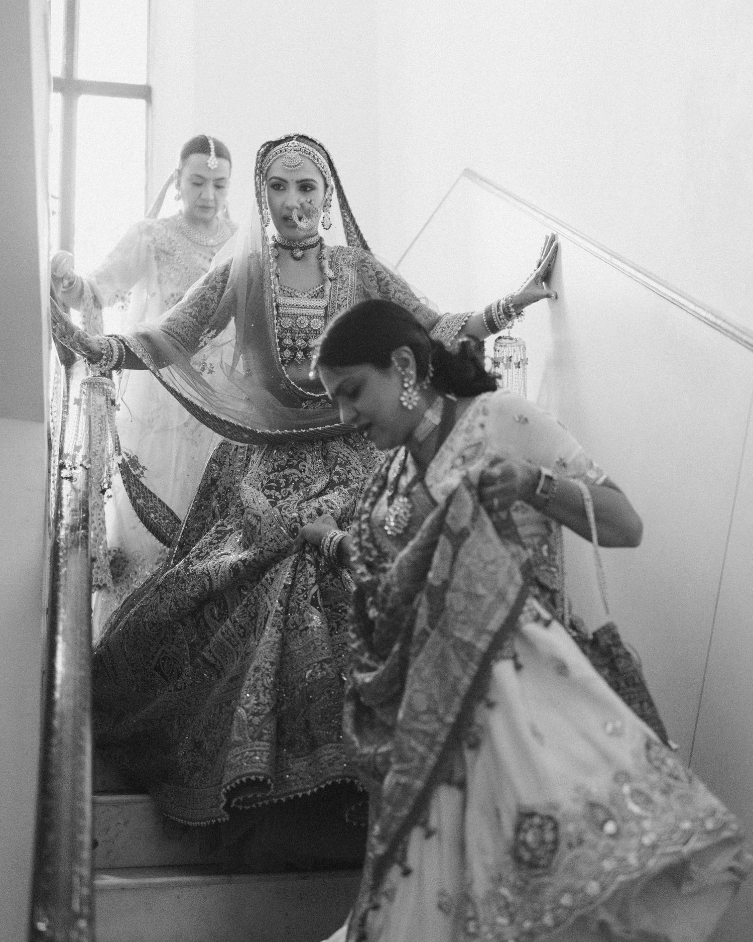 Black and white photo of Indian brides in traditional lehenga attire descending stairs during a wedding ceremony.