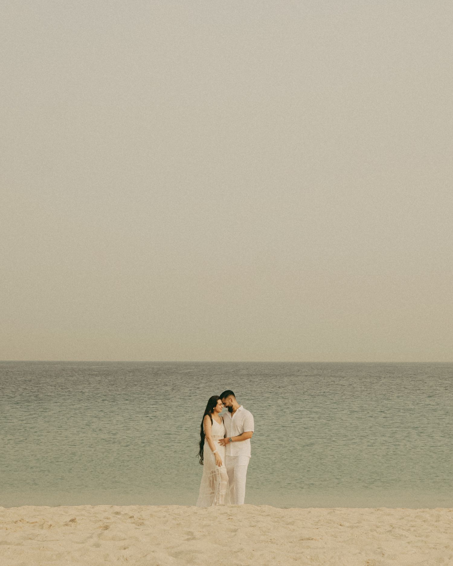 A couple in white outfits embrace romantically on a serene sandy beach with calm turquoise ocean waters behind them.