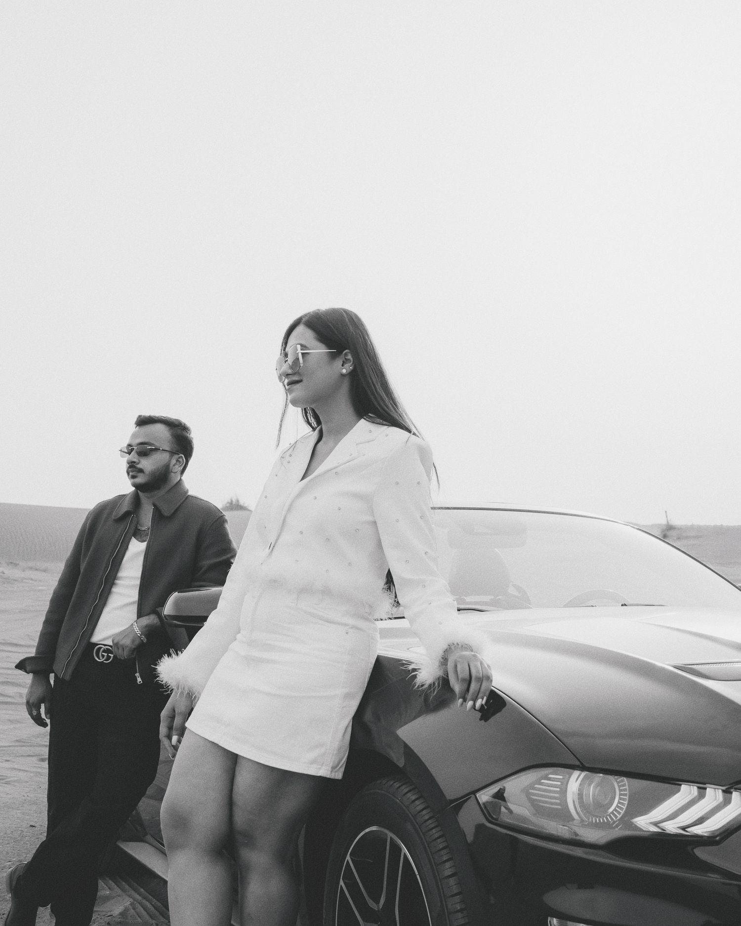 Black and white photo of a man and woman posing beside a luxury car outdoors.