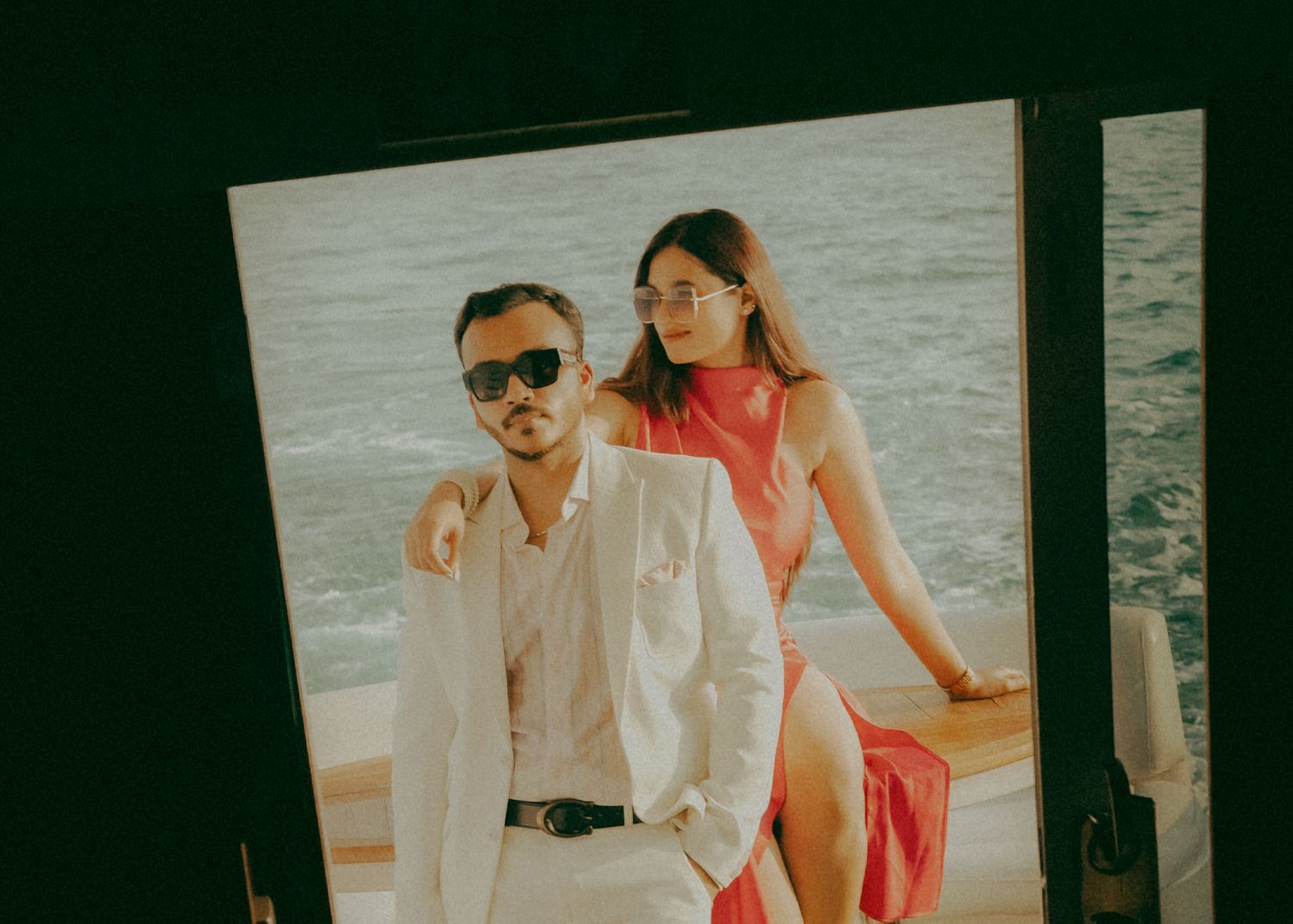 Stylish couple posing on a luxury yacht, man in white suit with sunglasses, woman in red dress by the ocean.