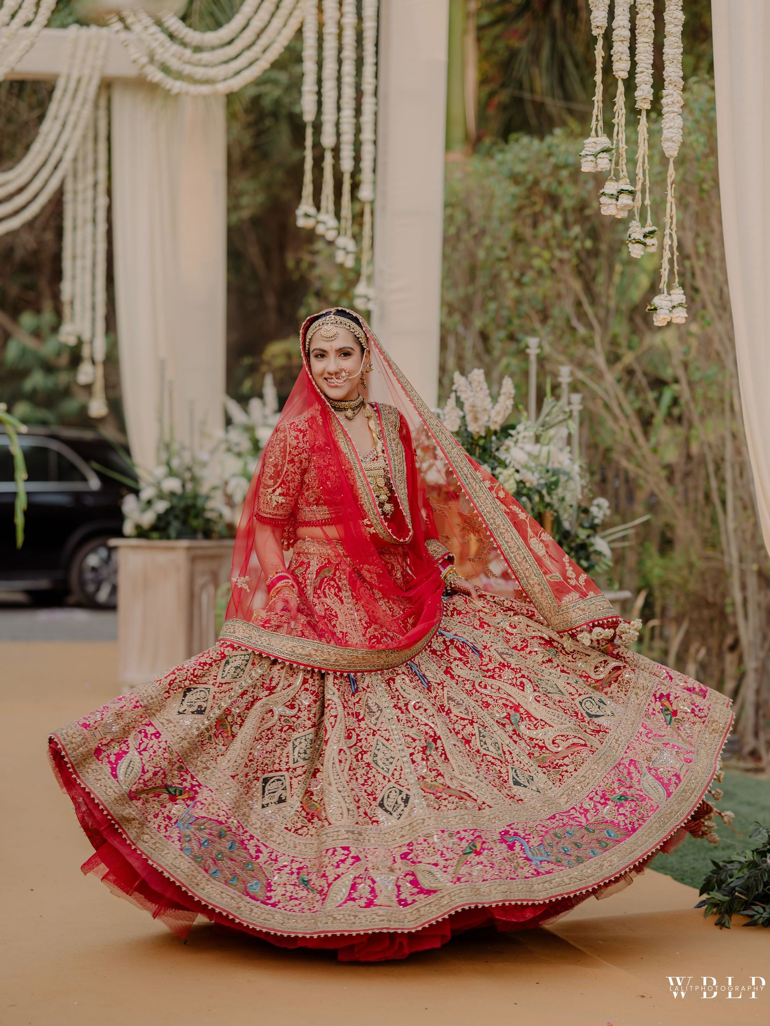 Bride twirling in red and gold embroidered bridal lehenga with dupatta at an outdoor wedding venue.