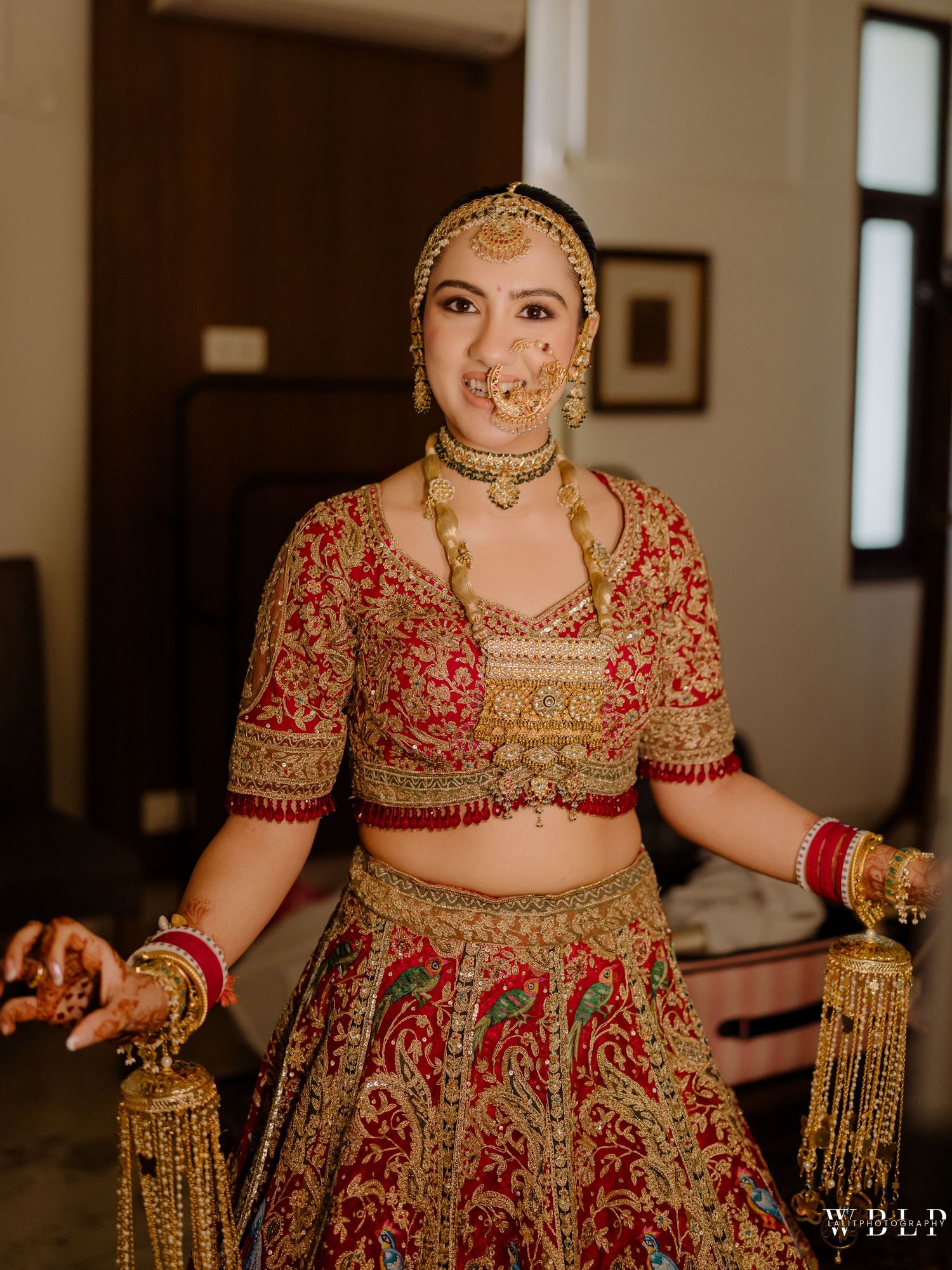 A bride in a red and gold embroidered lehenga with traditional jewelry, maang tikka, and nose ring poses indoors.