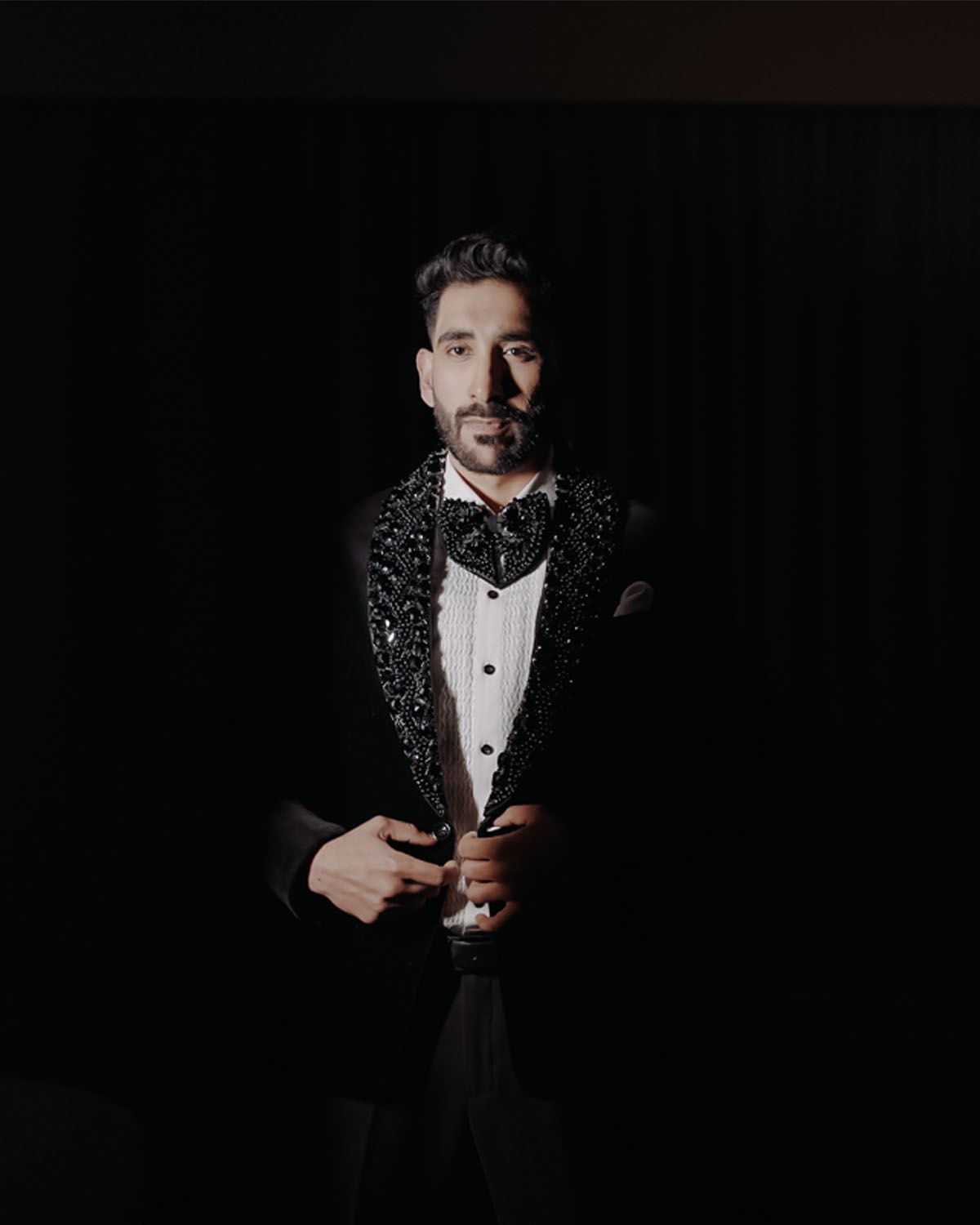 Dramatic portrait in sparkly black tuxedo jacket and bow tie against dark background.