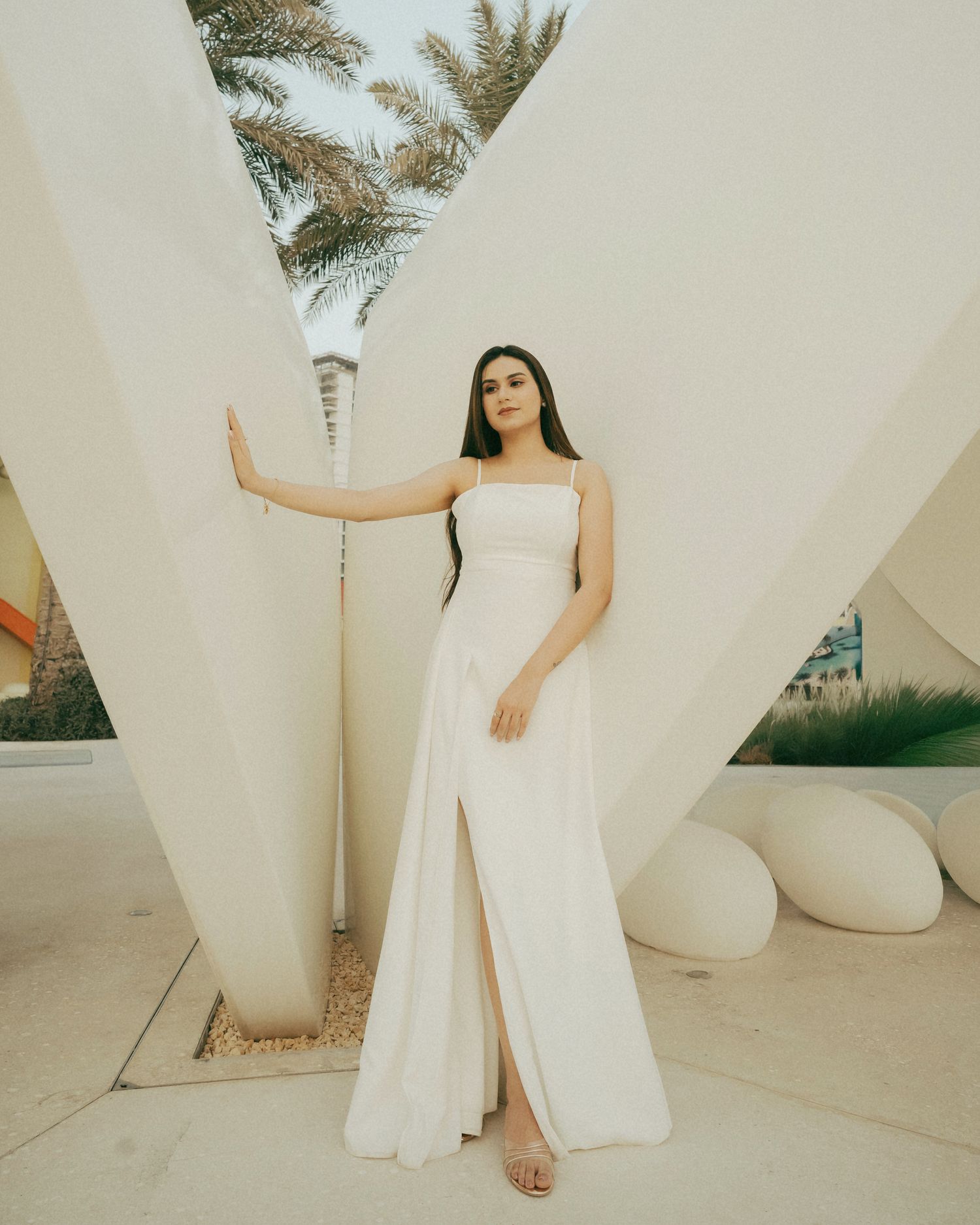 Woman in elegant white slit maxi dress posing gracefully between geometric white pillars with palm trees in background.