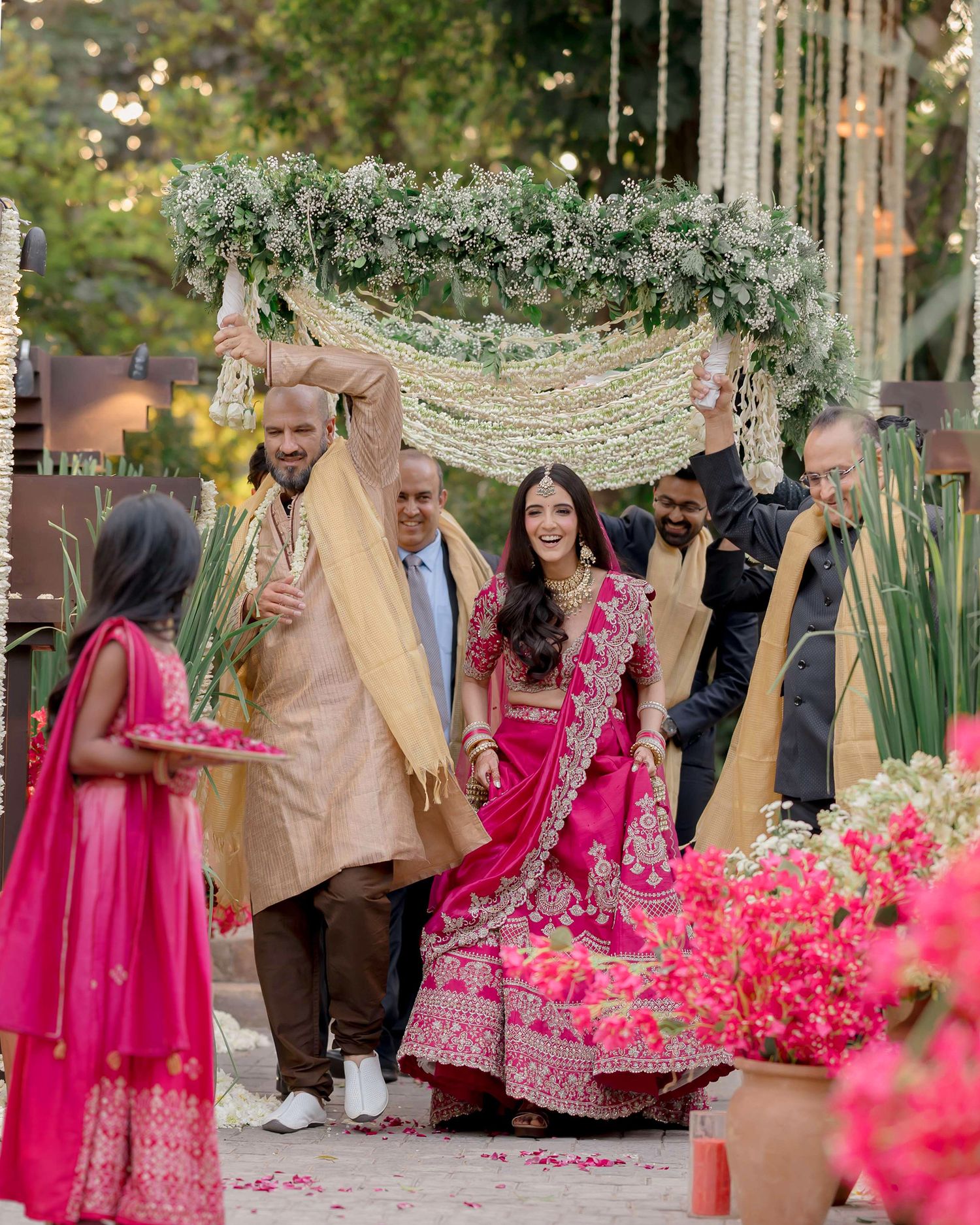 Bride in pink lehenga walks under floral canopy with family during traditional Indian wedding ceremony.