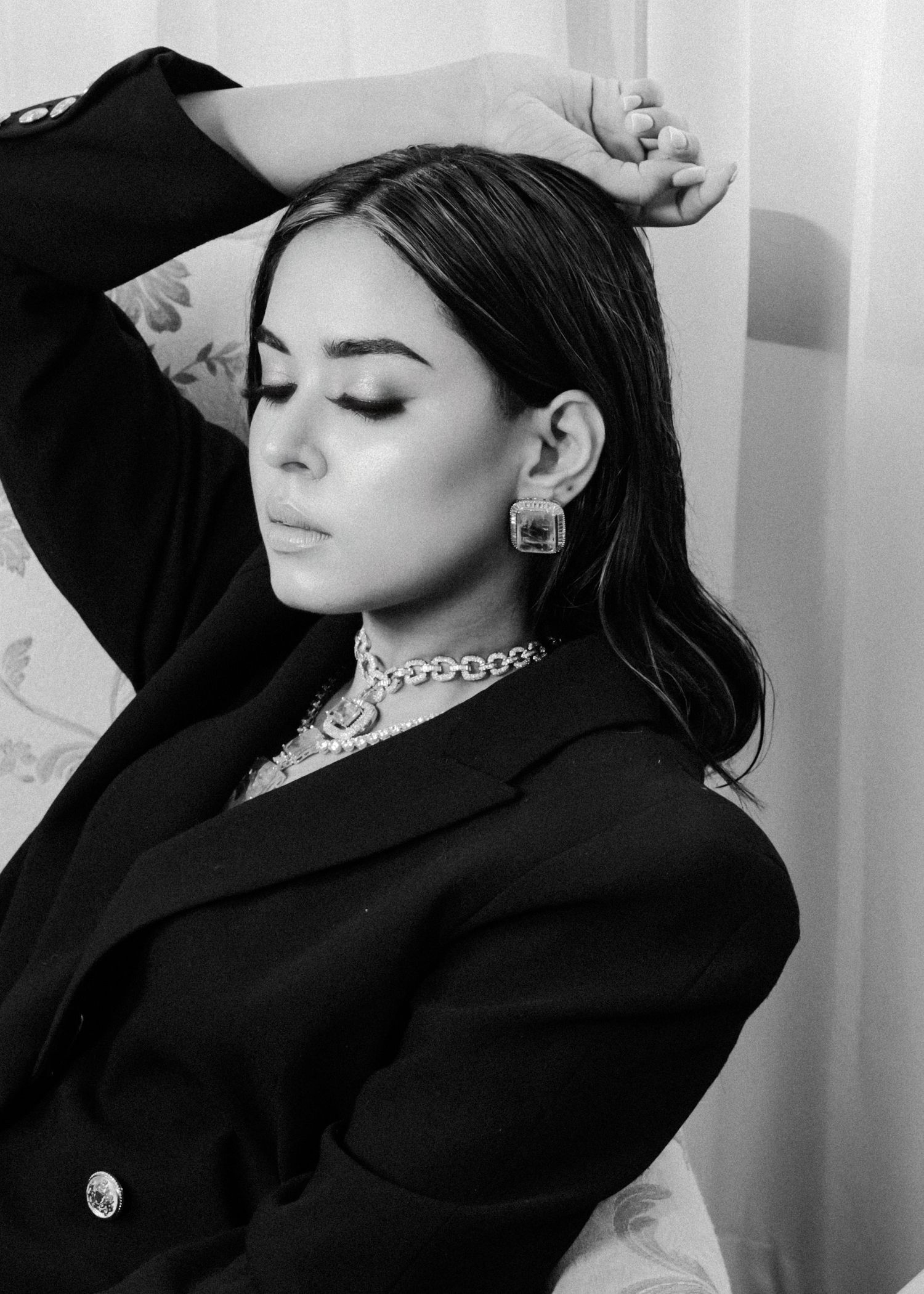 Black and white portrait of a person in a blazer wearing ornate jewelry and earrings with eyes closed against light background.