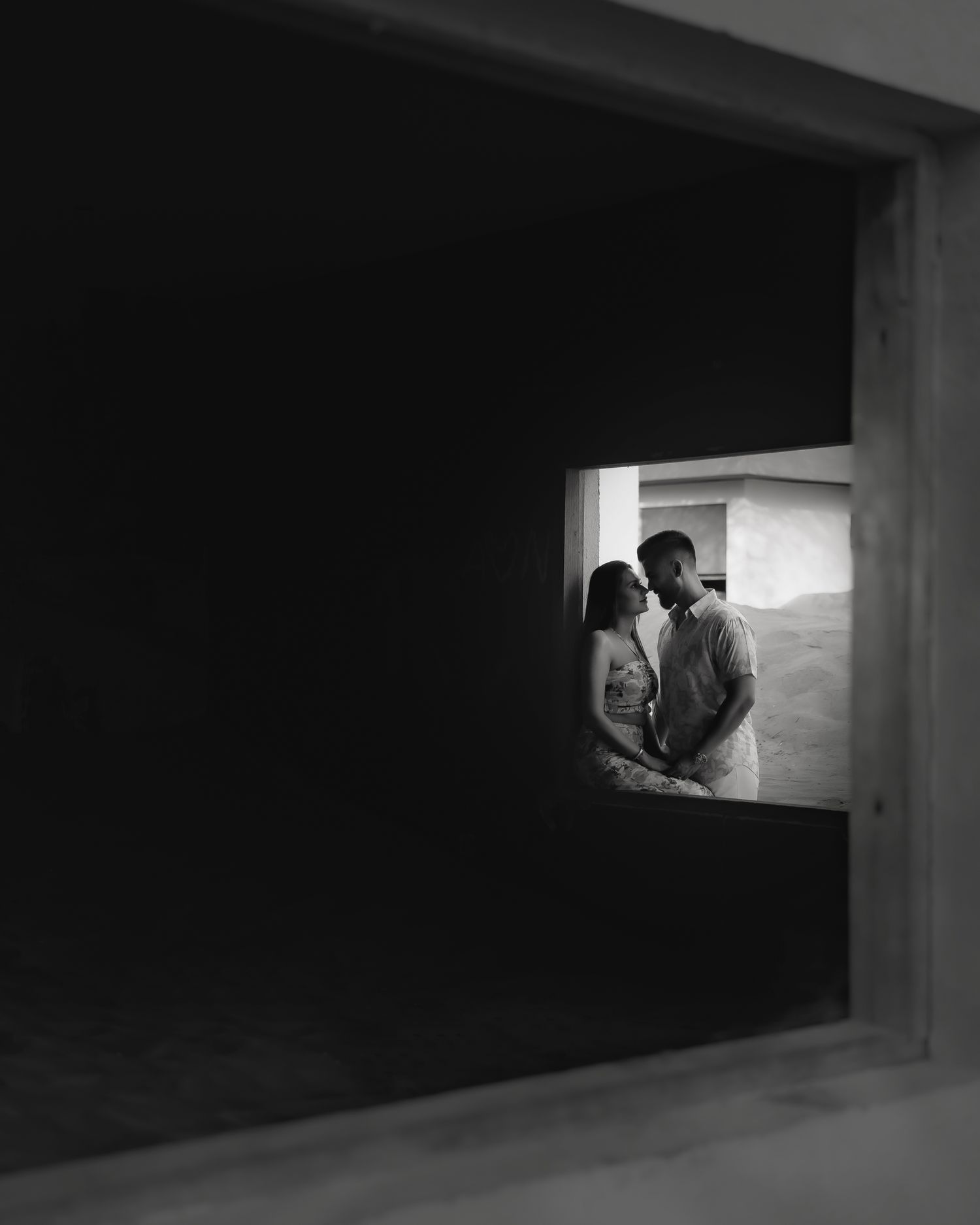 Black and white photo of a couple embracing in a small window opening, framed by dark walls.