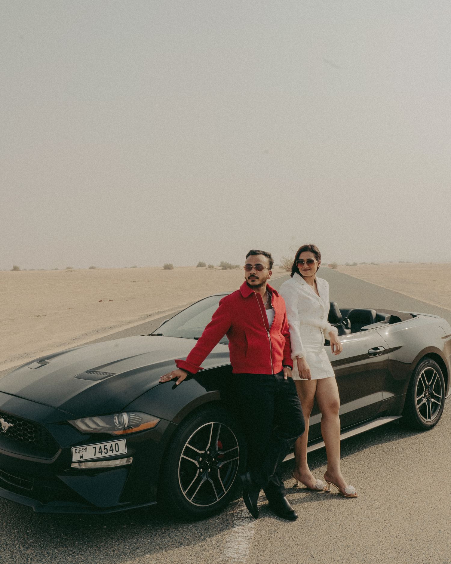 Couple posing by a black Ford Mustang convertible in a desert landscape, man in red jacket, woman in white dress.