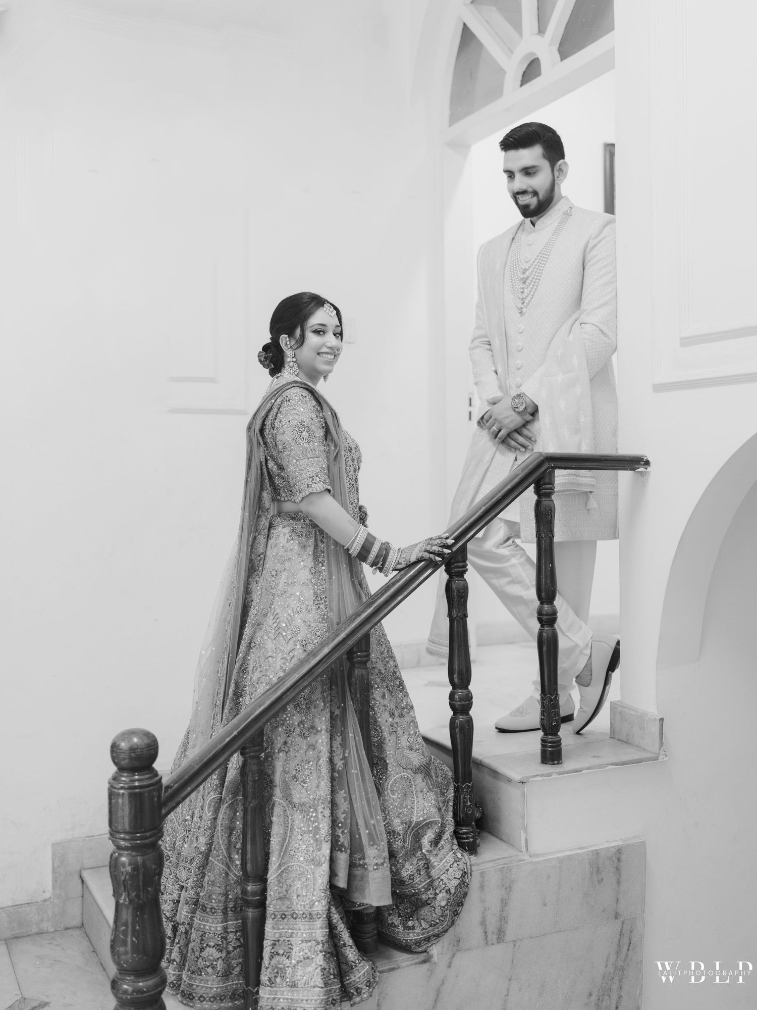 Elegant couple in traditional Indian wedding attire posing on white staircase in black and white photograph.
