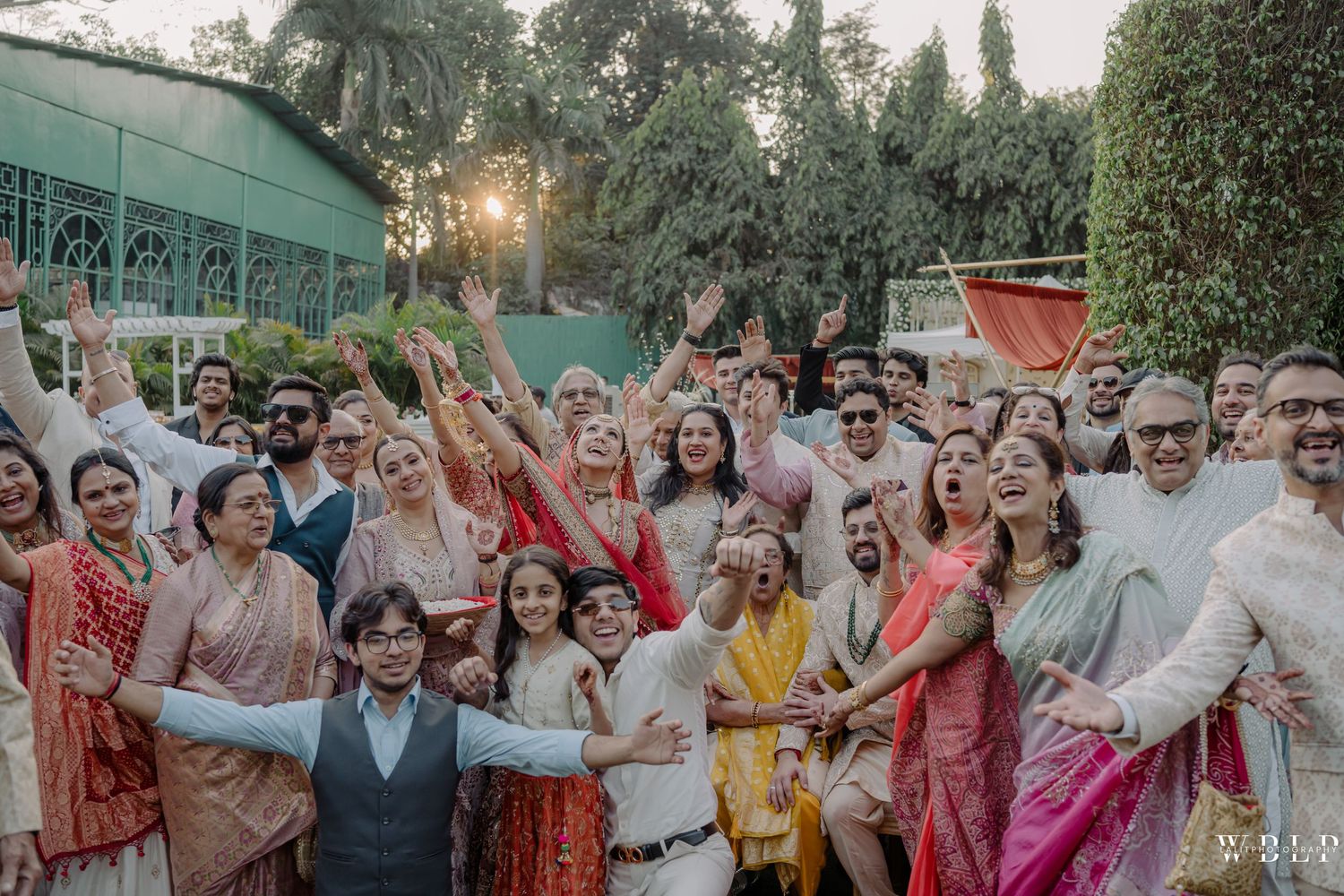 Large joyful Indian wedding group photo with guests in colorful traditional attire celebrating outdoors.
