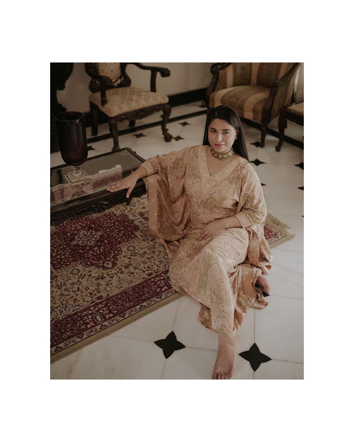 Person in beige dress seated on patterned floor beside antique furniture in classic colonial interior.