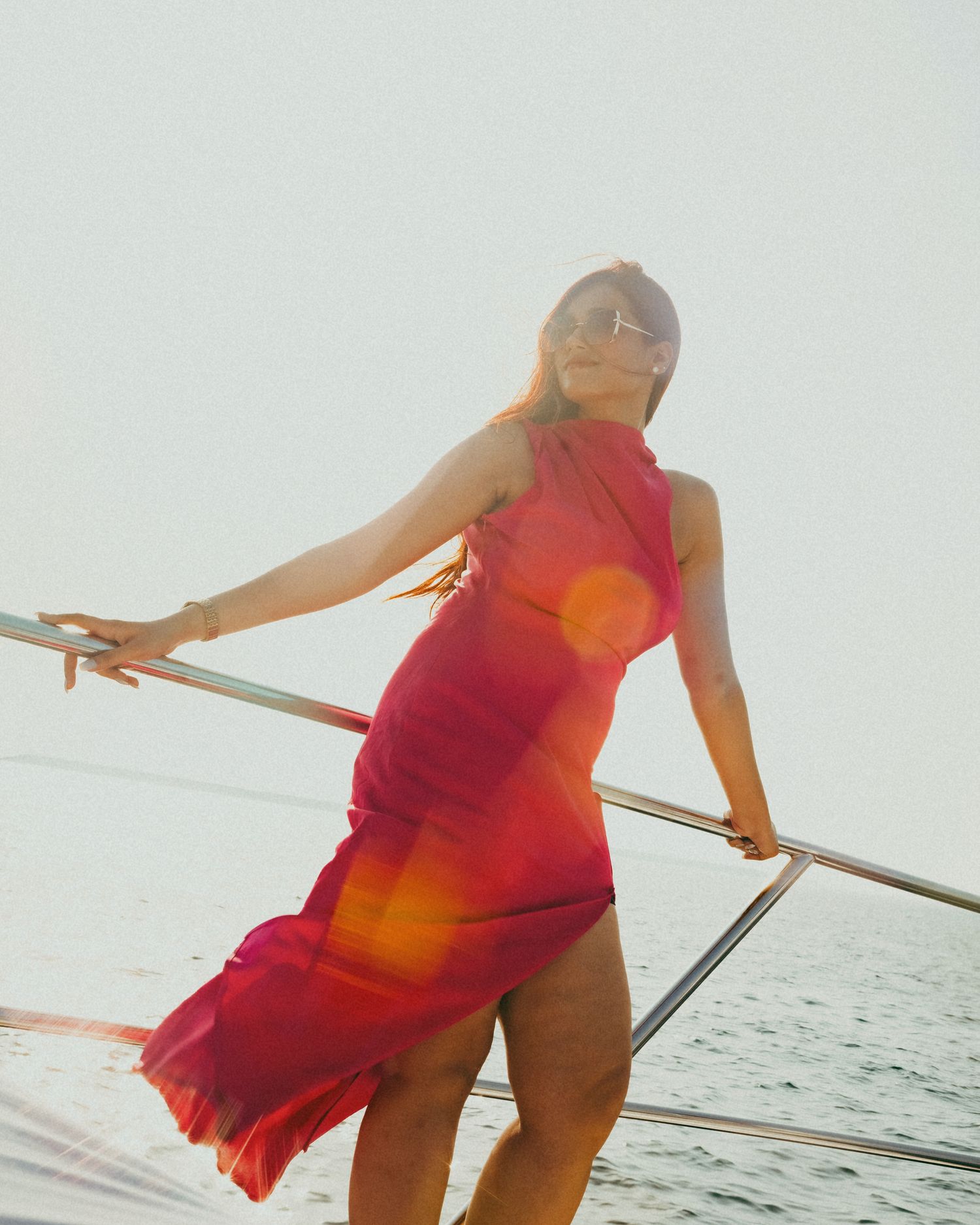 Woman in flowing red dress poses on yacht deck at sunset, hair blowing in breeze, wearing sunglasses.