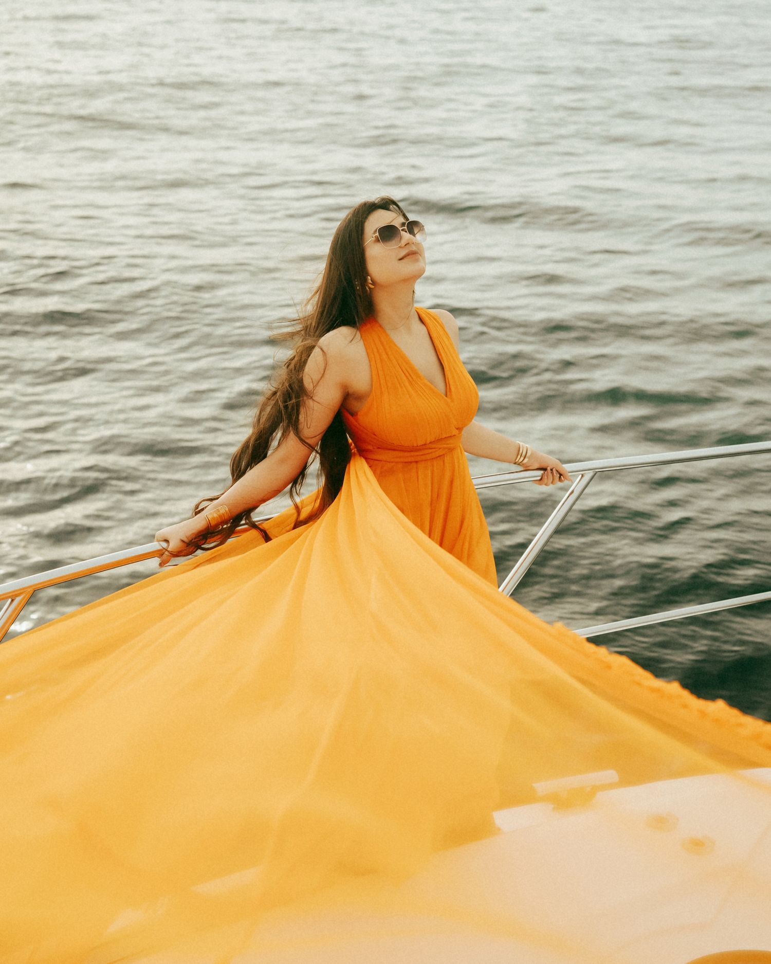 Woman in flowing orange halter dress poses on yacht bow, hair blowing in wind, wearing sunglasses with ocean backdrop.