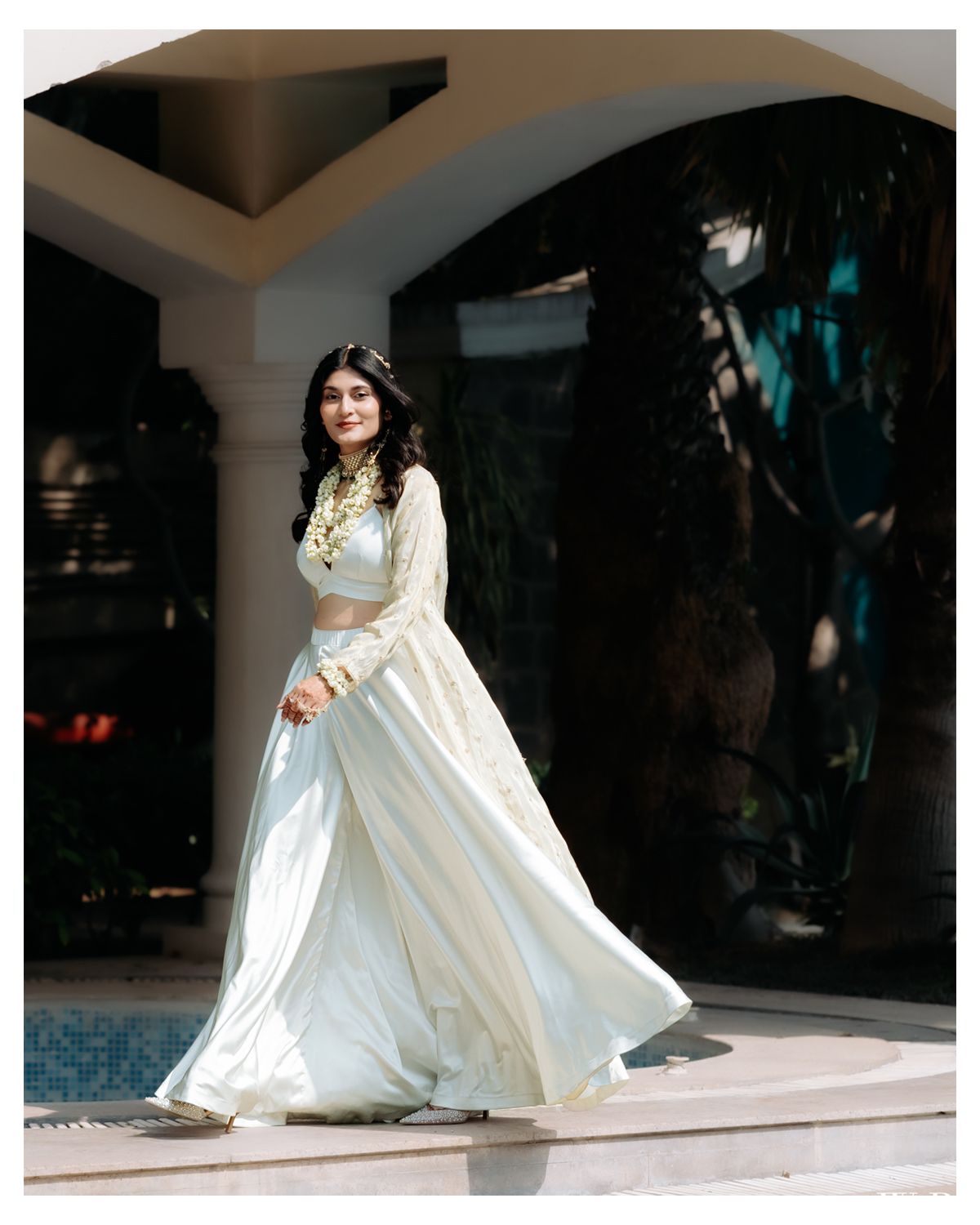 Bride in flowing white gown poses elegantly under an arched pavilion during a photoshoot.