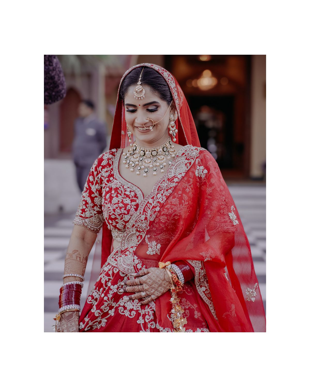 Bride in ornate red lehenga with detailed jewelry and embroidery smiles during traditional Indian wedding ceremony.