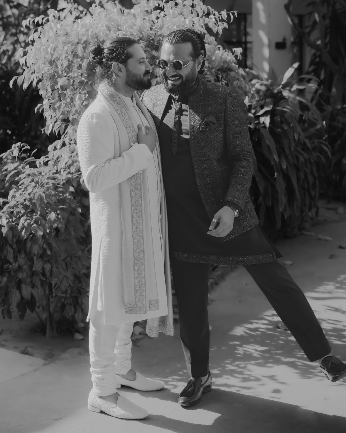 Black and white photo of two people in contrasting outfits sharing a candid moment outdoors in dappled sunlight.