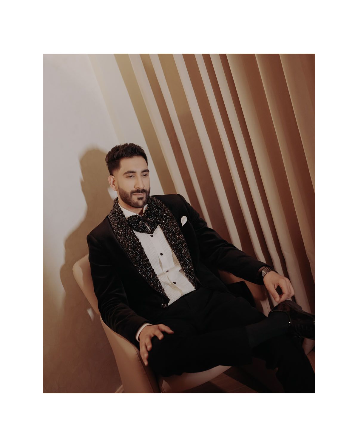 Stylish portrait in glittering black tuxedo while seated against textured wall.