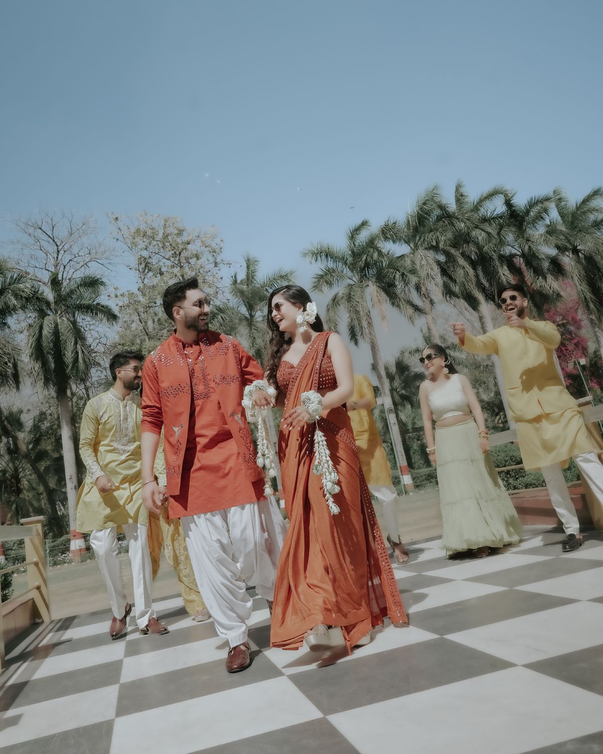 Wedding party in traditional Indian attire walking on a checkered floor with palm trees in the background.