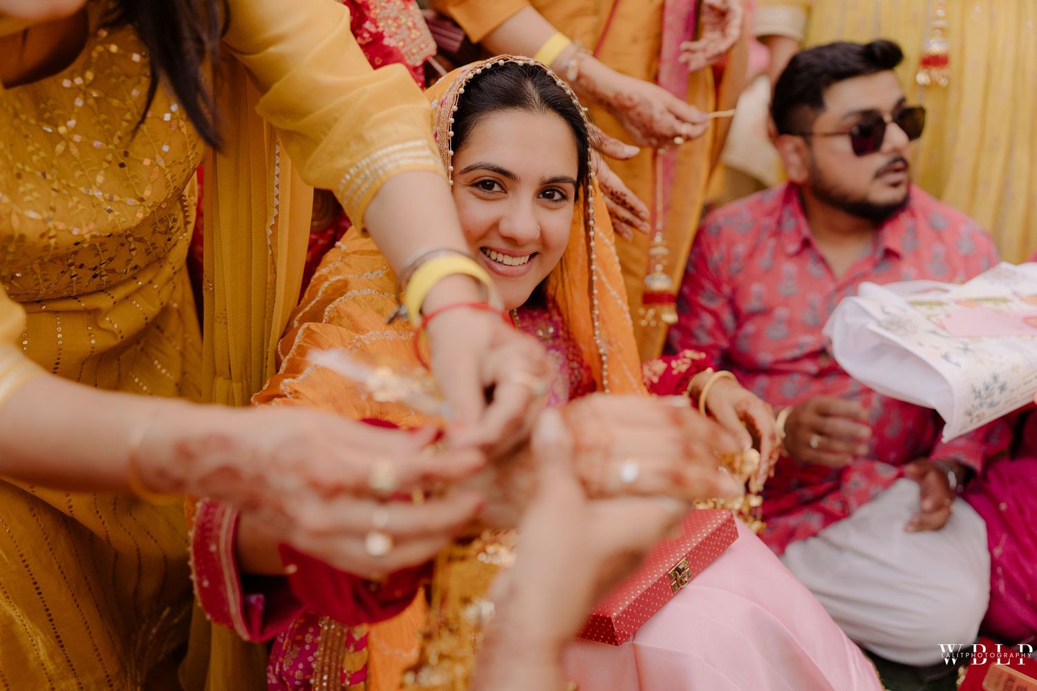 Smiling bride in pink and gold attire during a traditional Indian wedding ceremony with guests in vibrant outfits.