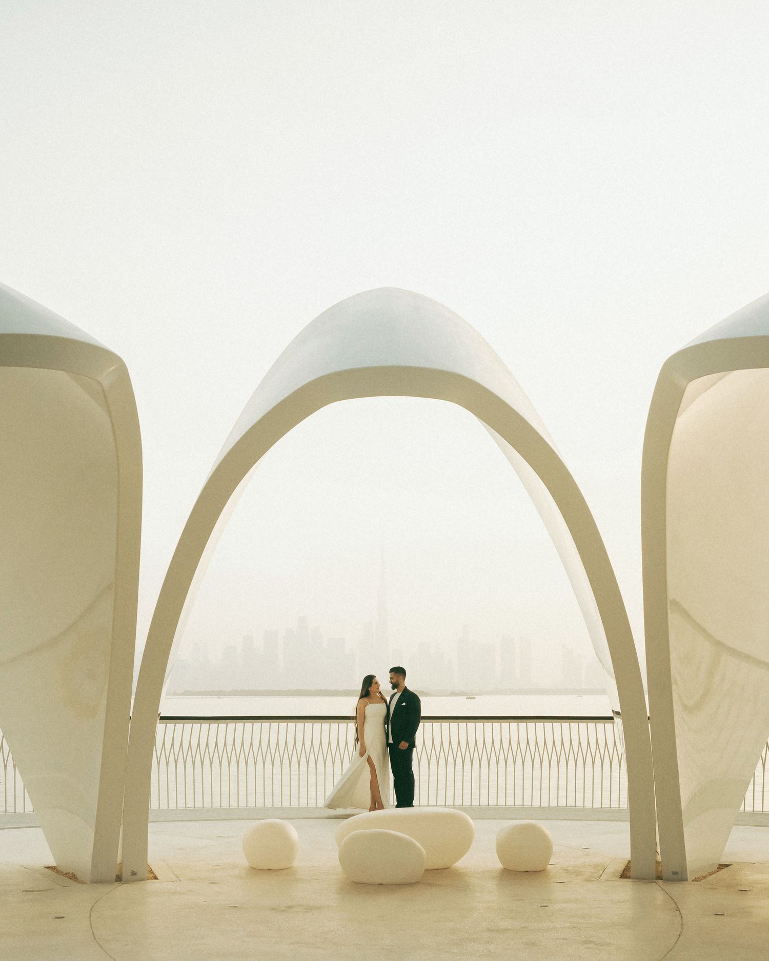 Couple poses under white architectural arches with Dubai skyline backdrop at a modern waterfront wedding venue.