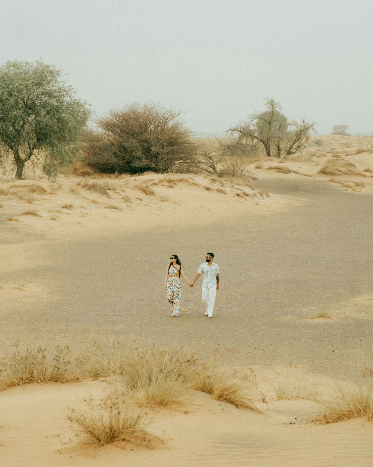 A couple walks hand in hand through golden desert sand dunes with sparse trees in the background.