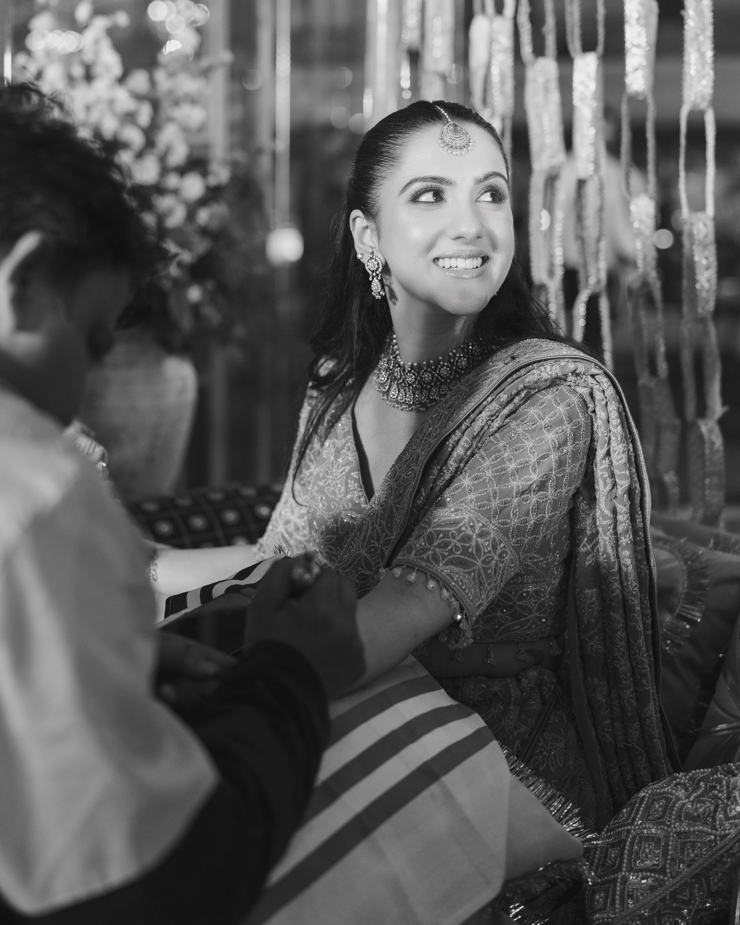 Black and white photo of a smiling woman in traditional Indian bridal attire with jewelry at a wedding ceremony.