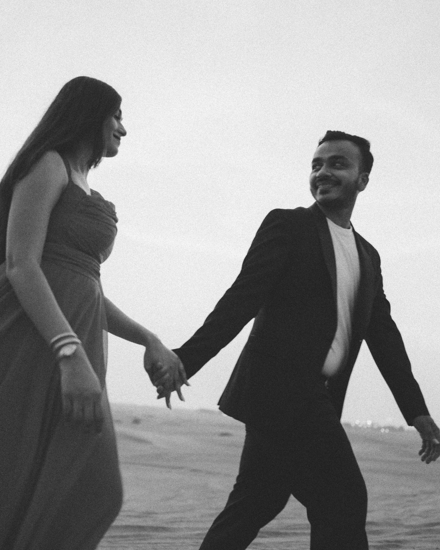 Black and white photo of a smiling couple holding hands and walking on a beach, woman in elegant gown, man in suit.
