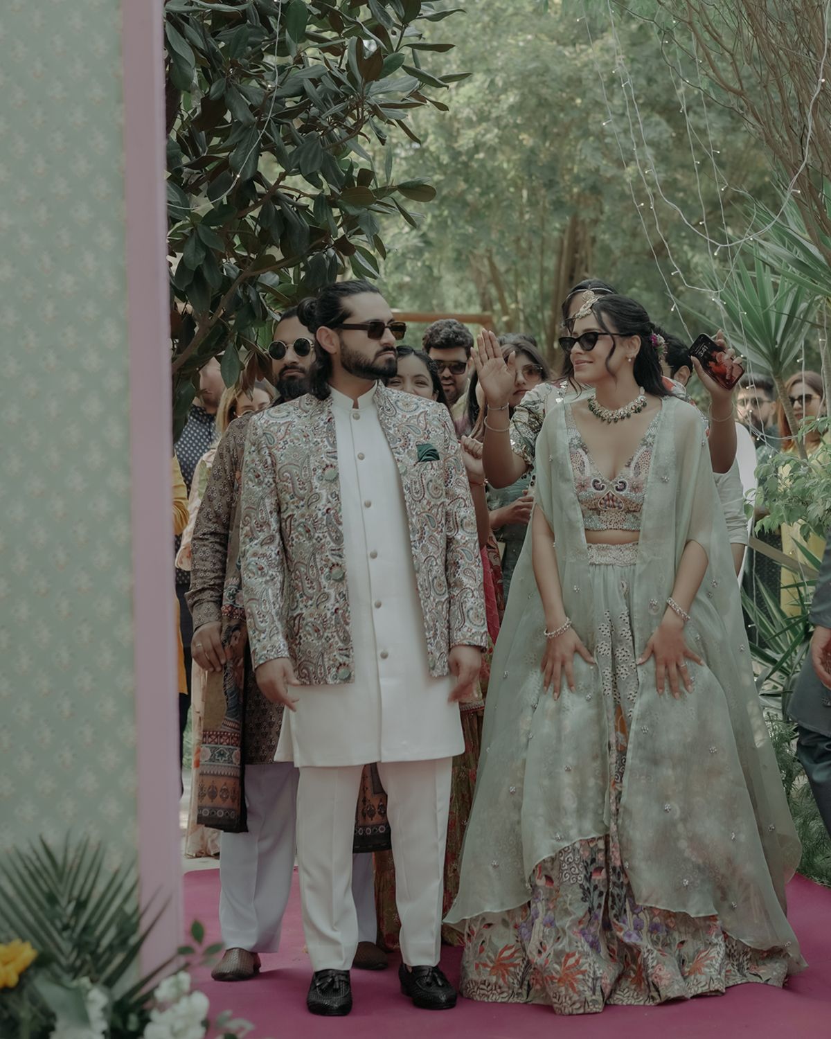 A couple in traditional Indian wedding attire with gold embroidery stand together in a lush garden setting.