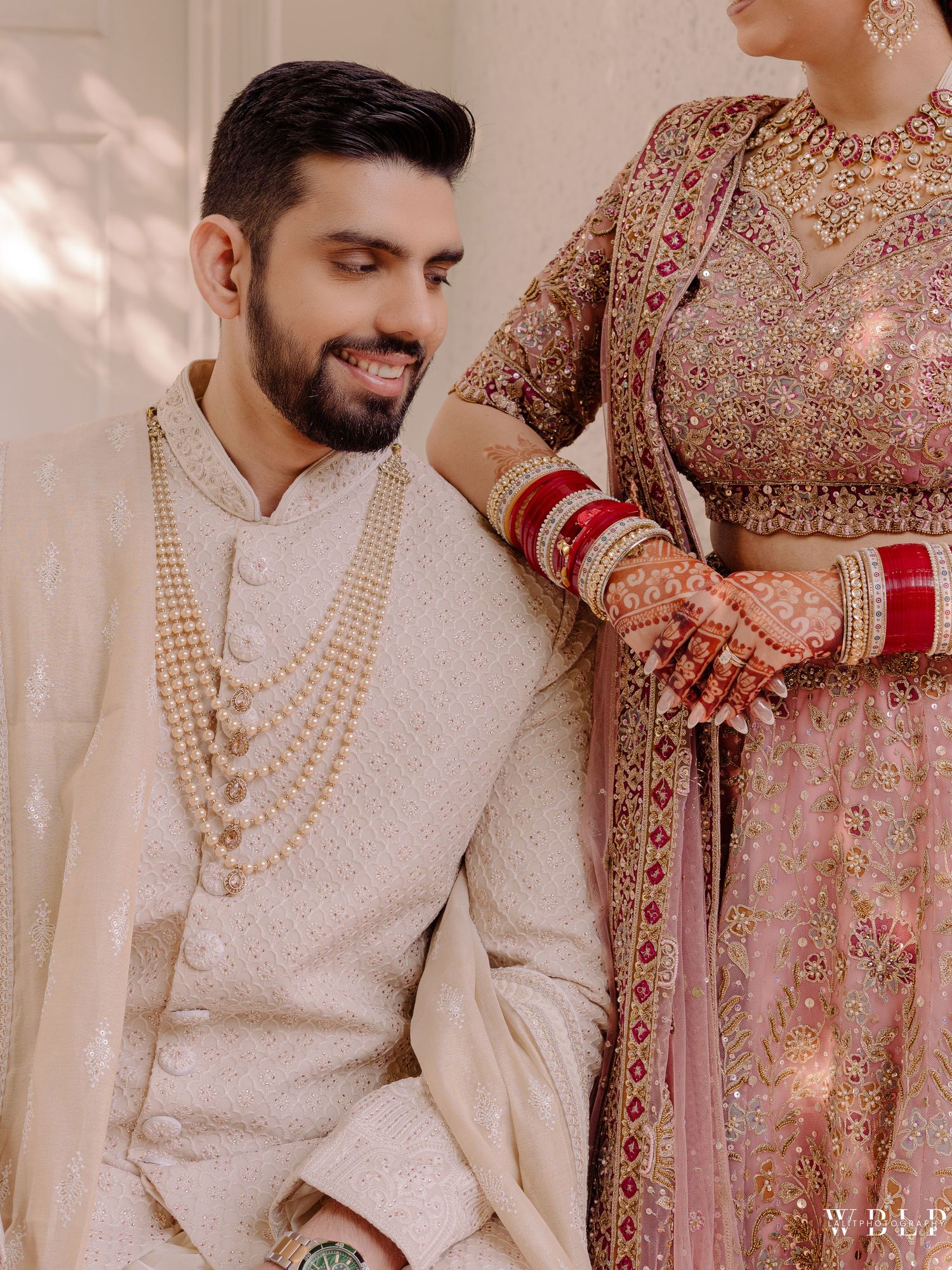 Indian groom in cream sherwani with pearl mala smiles at bride in pink embroidered lehenga with red bangles and mehndi.