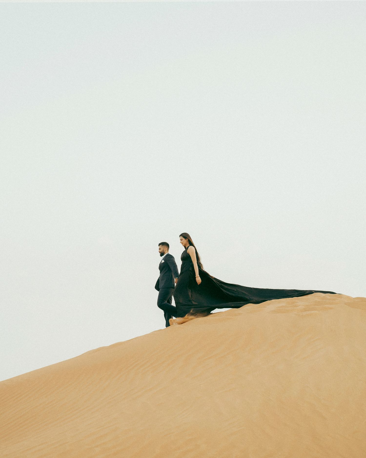 Couple in formal attire walks atop a golden sand dune, woman's black gown trailing dramatically in the desert breeze.