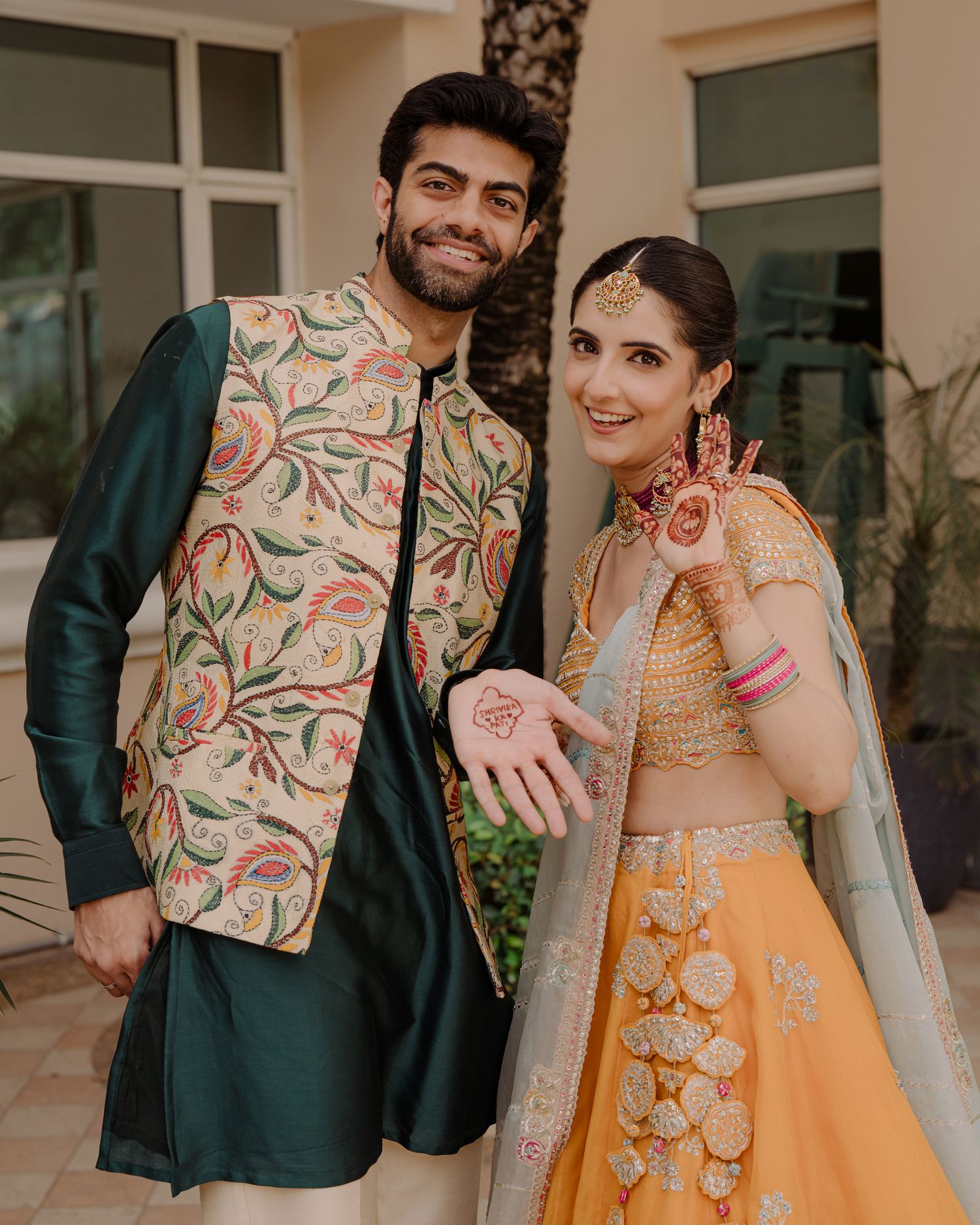 Couple in traditional Indian attire, man in green kurta with floral vest, woman in yellow lehenga showing mehndi.