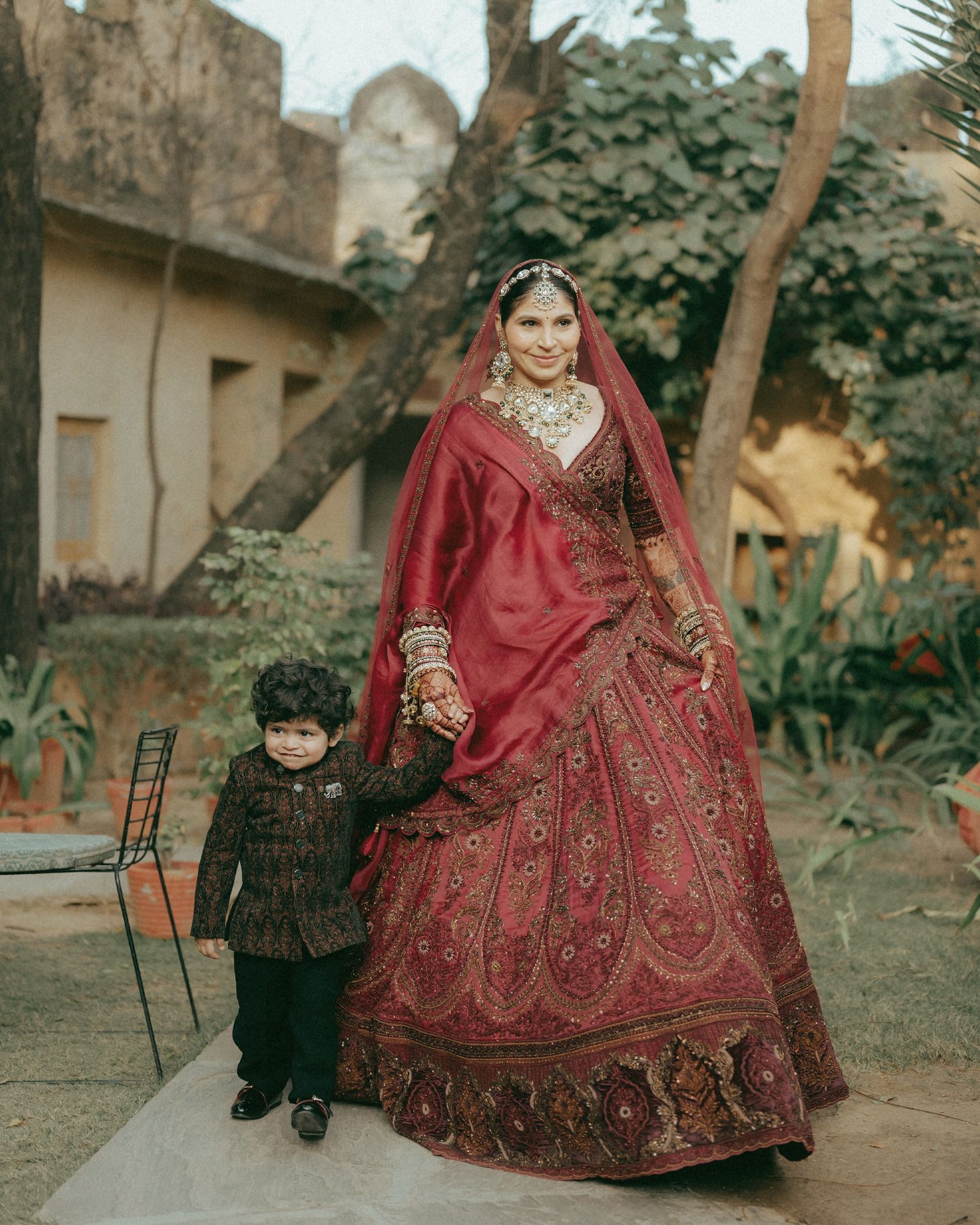 Indian bride in red lehenga with gold jewelry holds hands with young boy in traditional black outfit at outdoor venue.