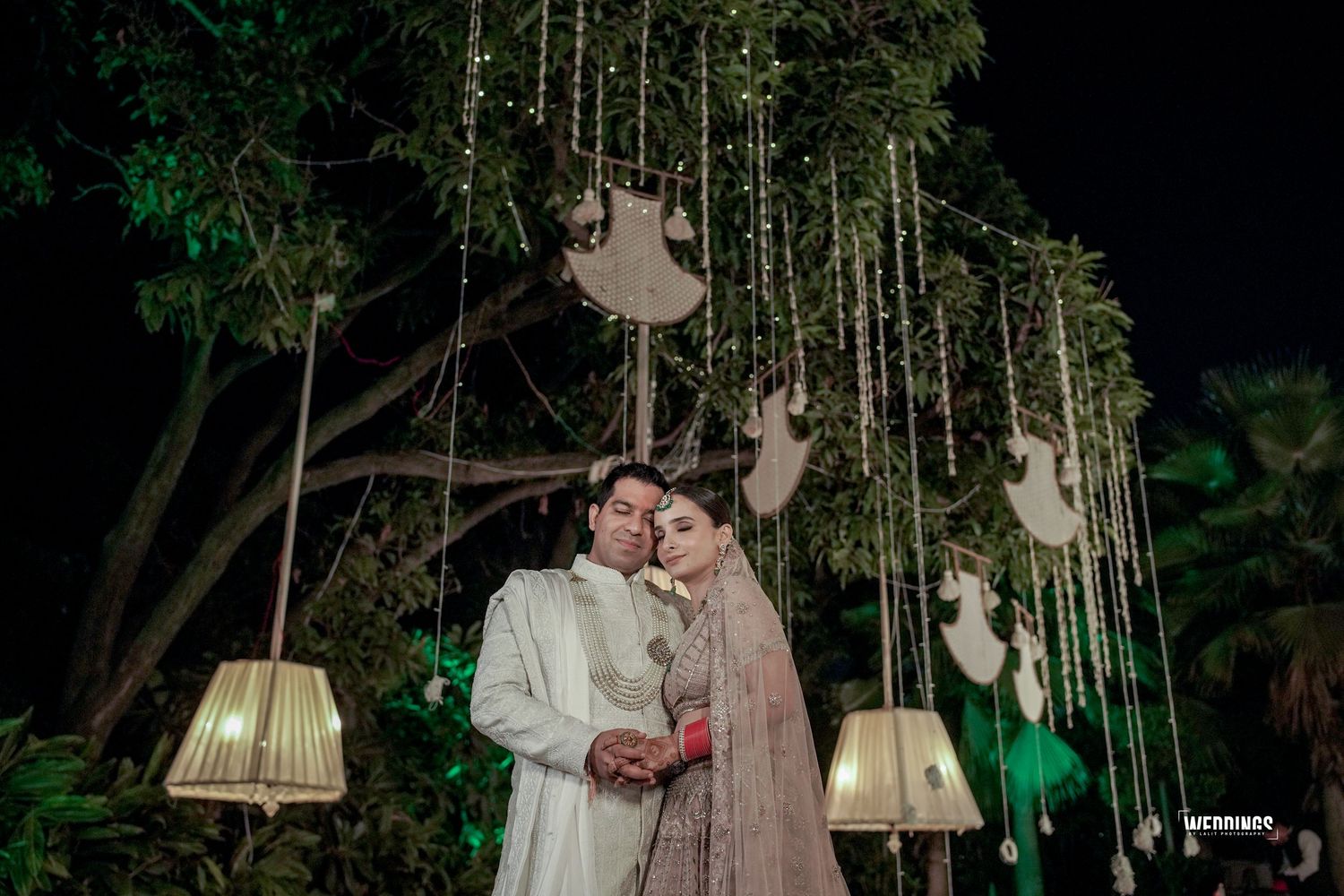 A romantic nighttime wedding reception decorated with hanging lamps and ethereal lights in an outdoor garden setting.