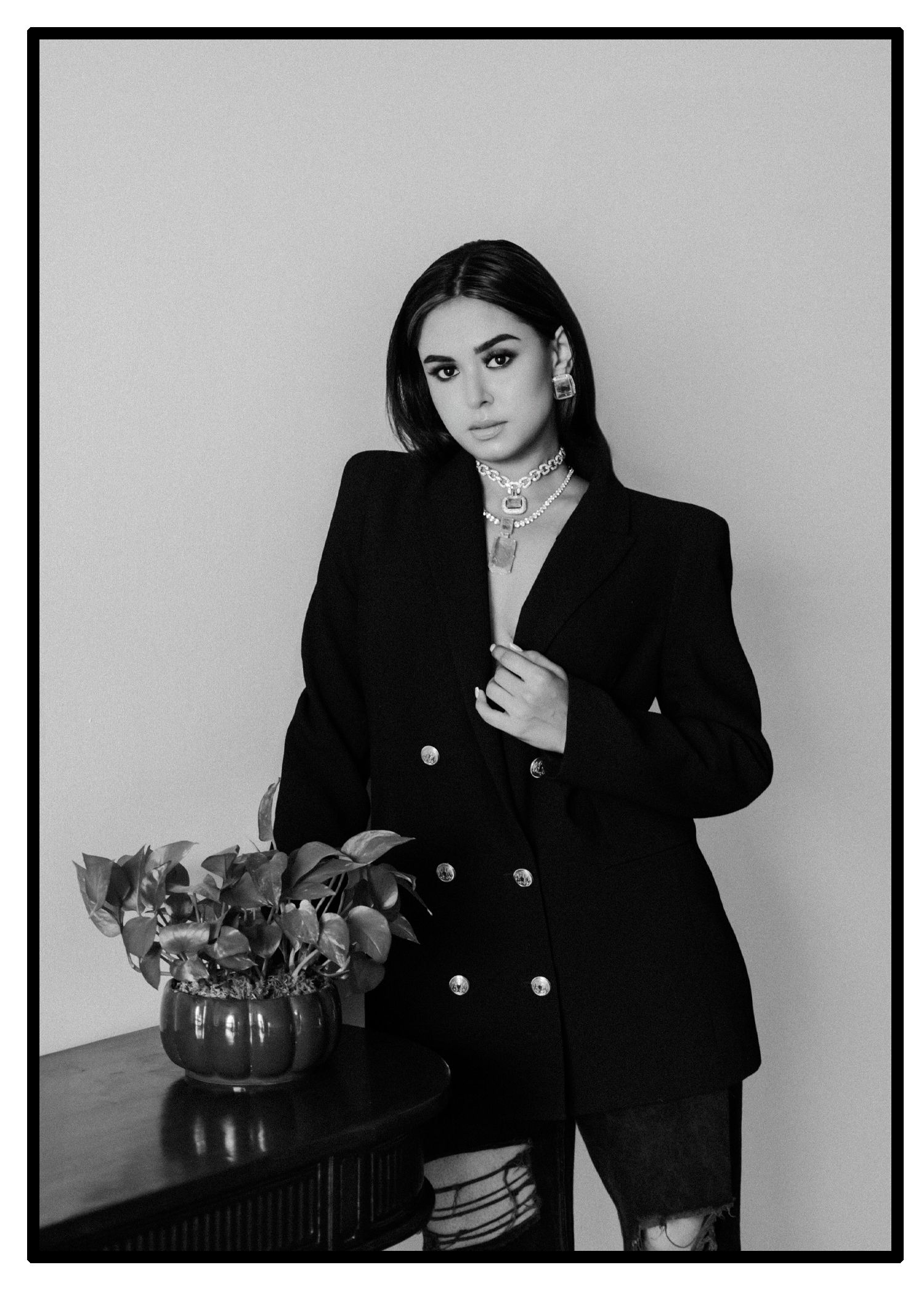Black and white editorial portrait featuring a formal blazer and layered necklaces beside a potted plant.