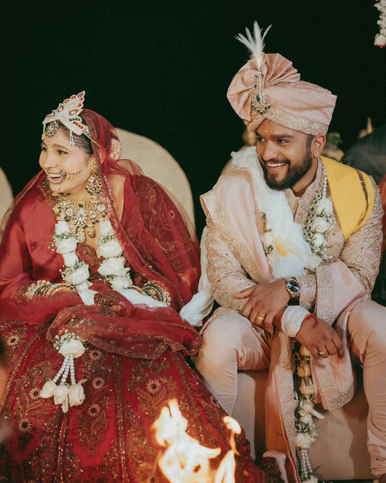 A bride in red lehenga and groom in cream sherwani smile during traditional Indian wedding ceremony with sacred fire.